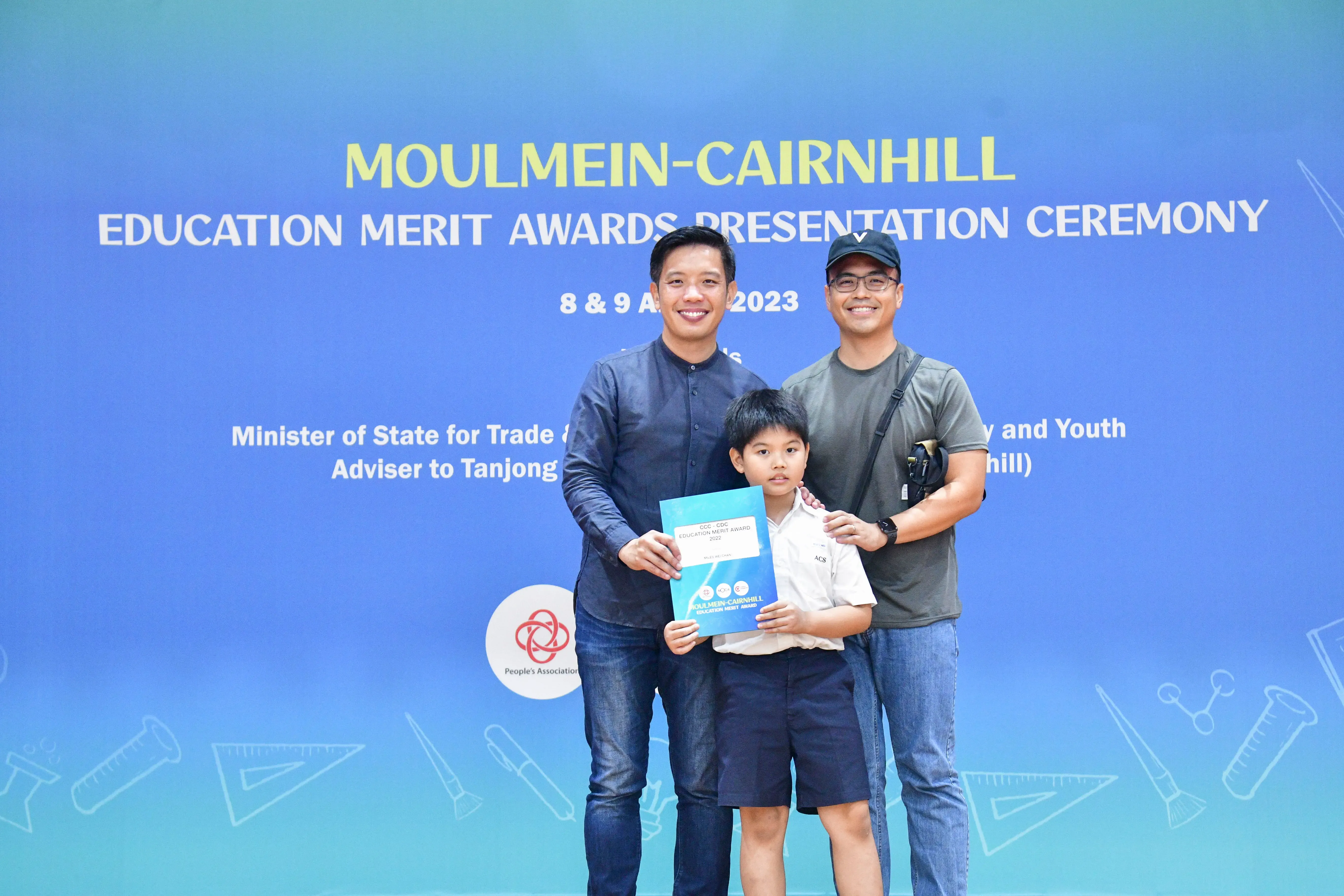 Two men and a young boy holding an award certificate pose in front of a Moulmein-Cairnhill Education Merit Awards banner.