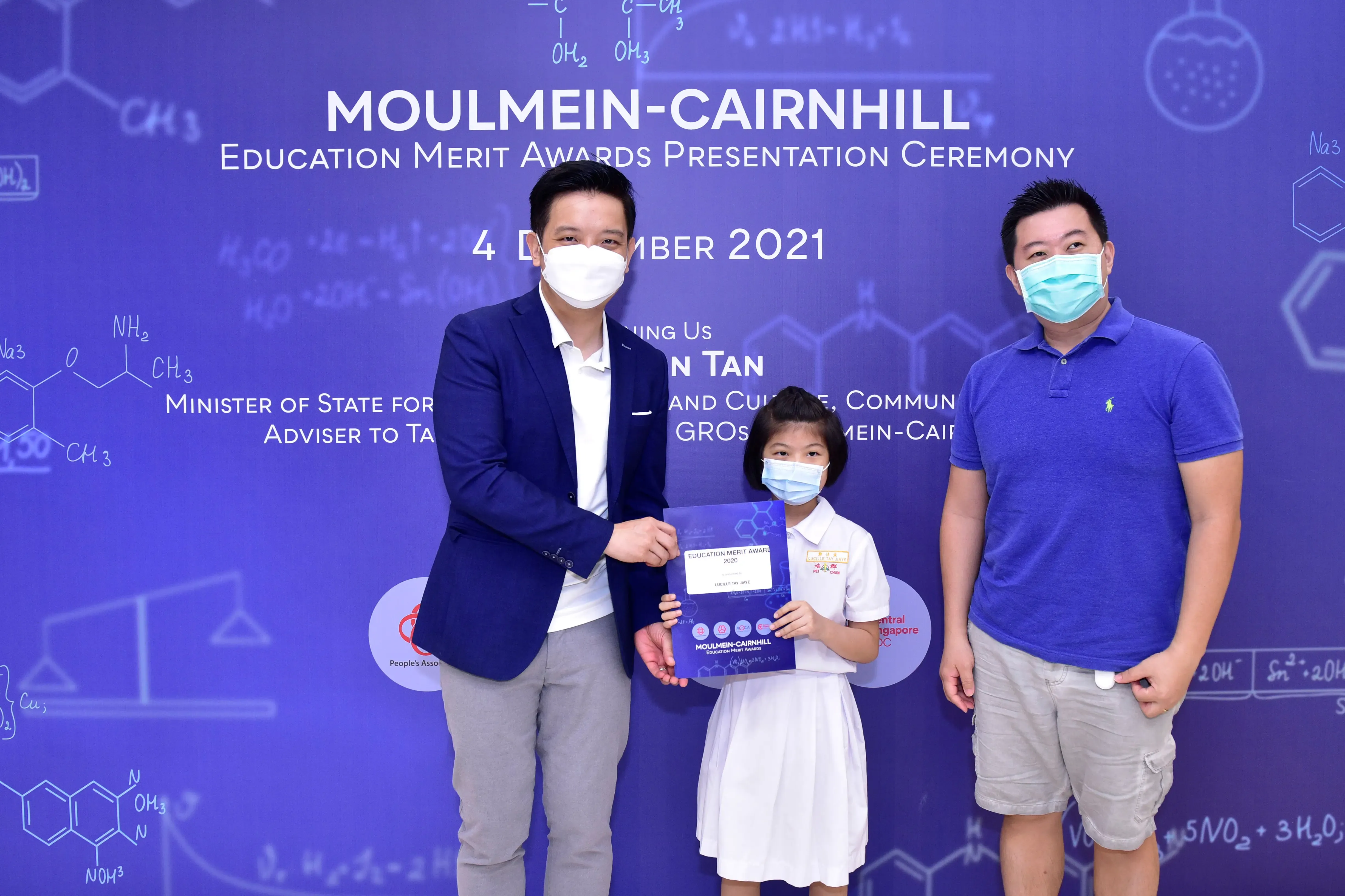 Two masked men and a young girl holding a certificate at the Moulmein-Cairnhill Education Merit Awards 2021.