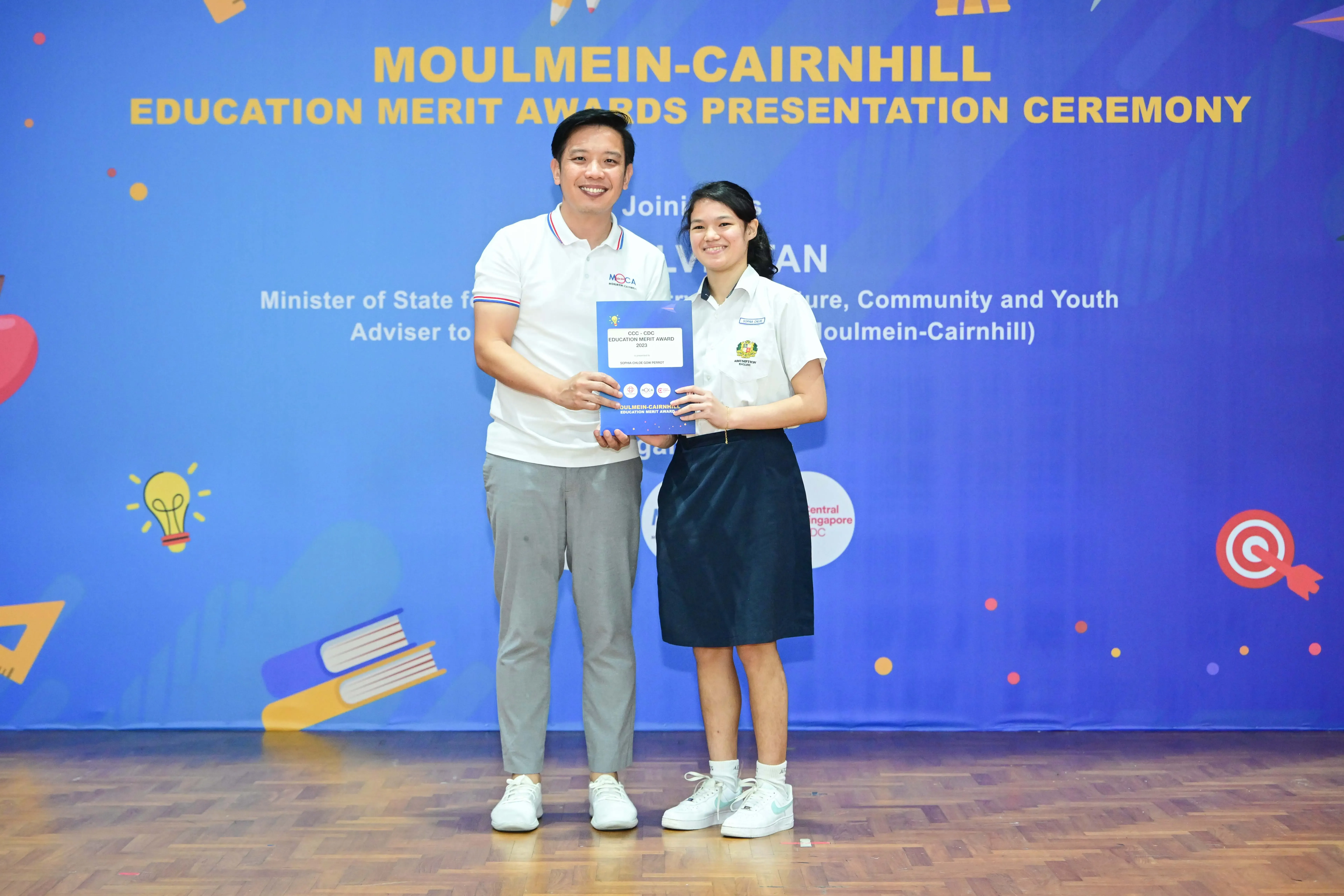 A man and a female student hold an award certificate at the Moulmein-Cairnhill Education Merit Awards Presentation Ceremony.