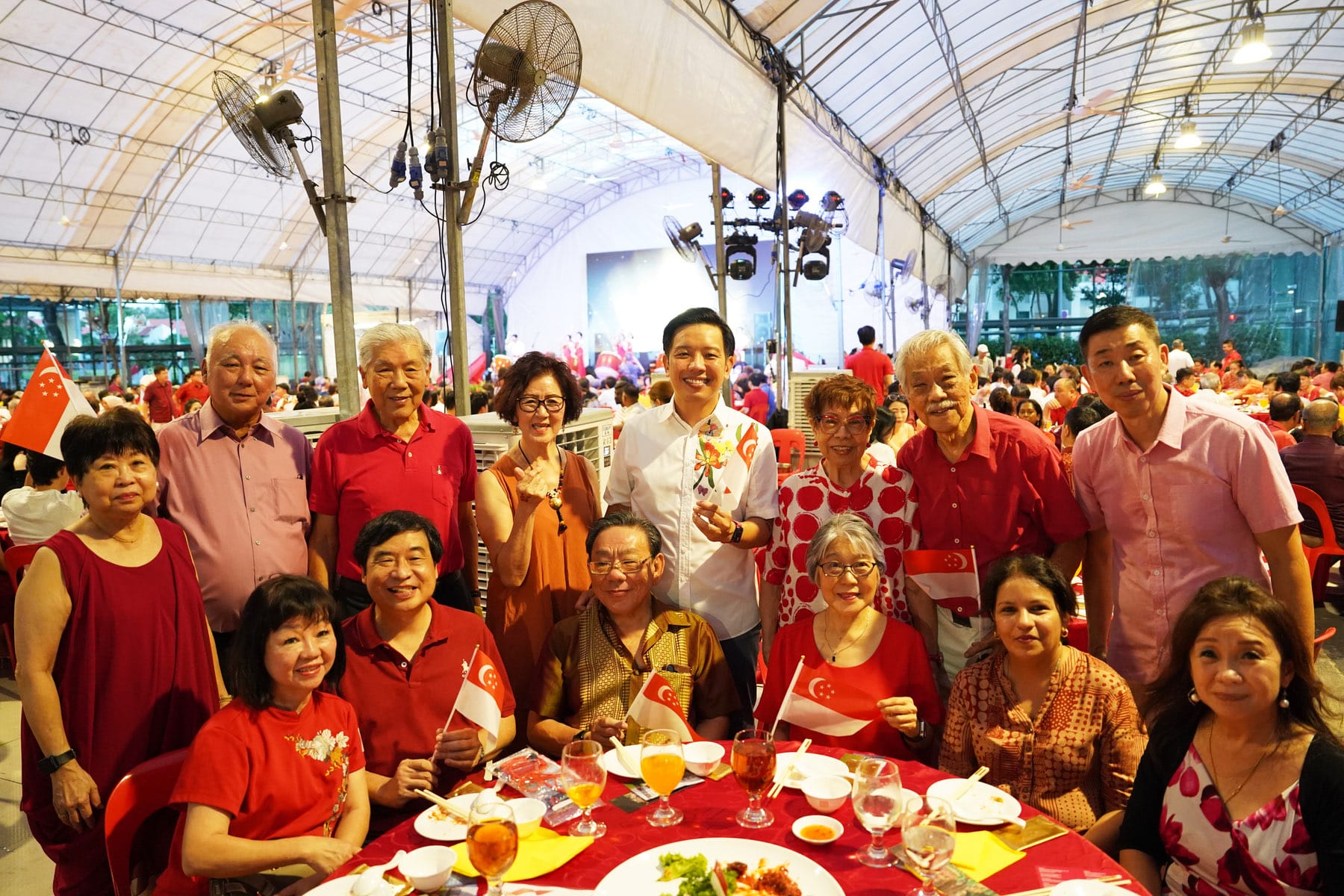 A large group of people in red outfits pose at a festive dinner event, holding small Singapore flags under a white tent.