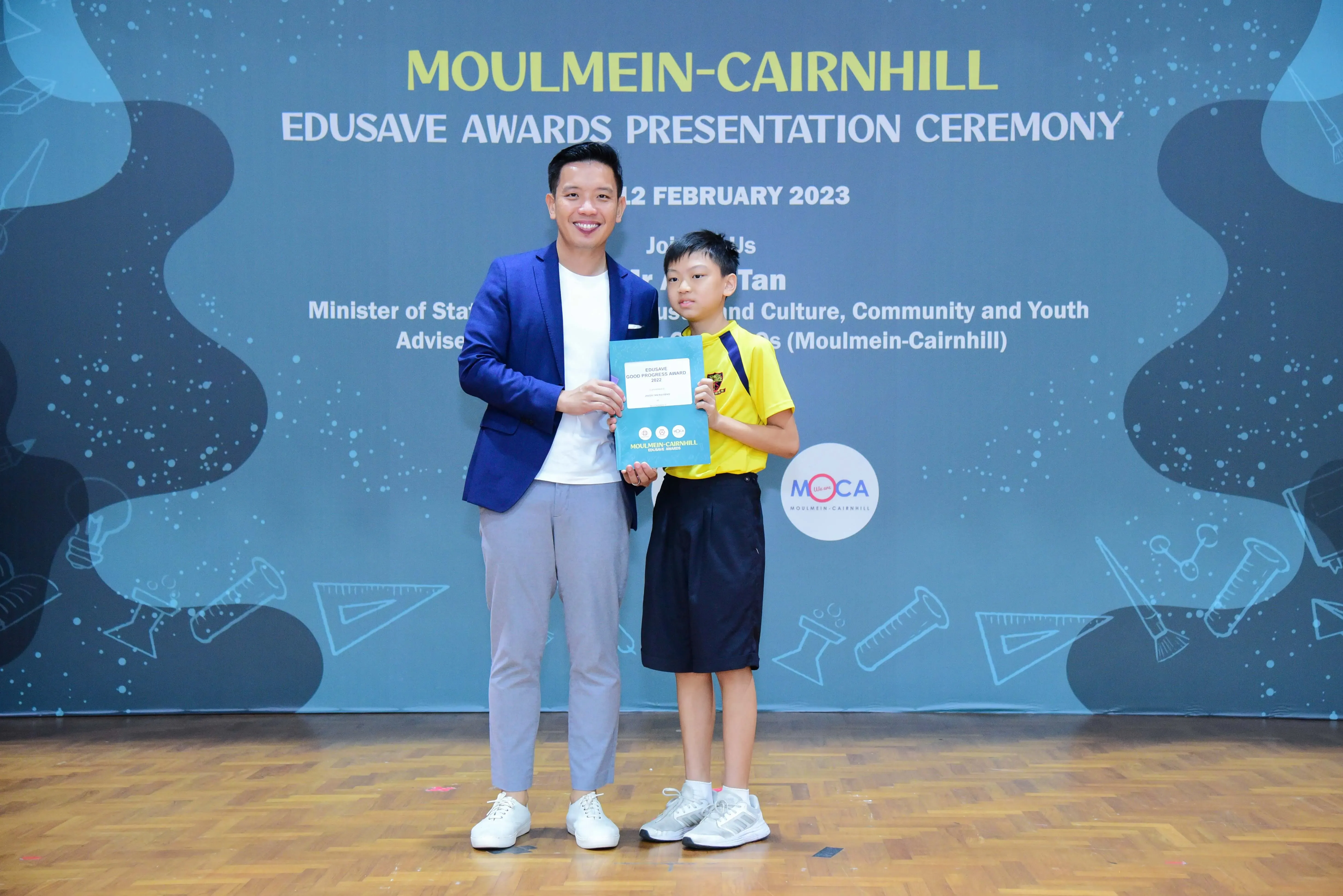 An adult in a blue blazer and a young boy in a yellow shirt hold a certificate at the Moulmein-Cairnhill Edusave Awards 2023.