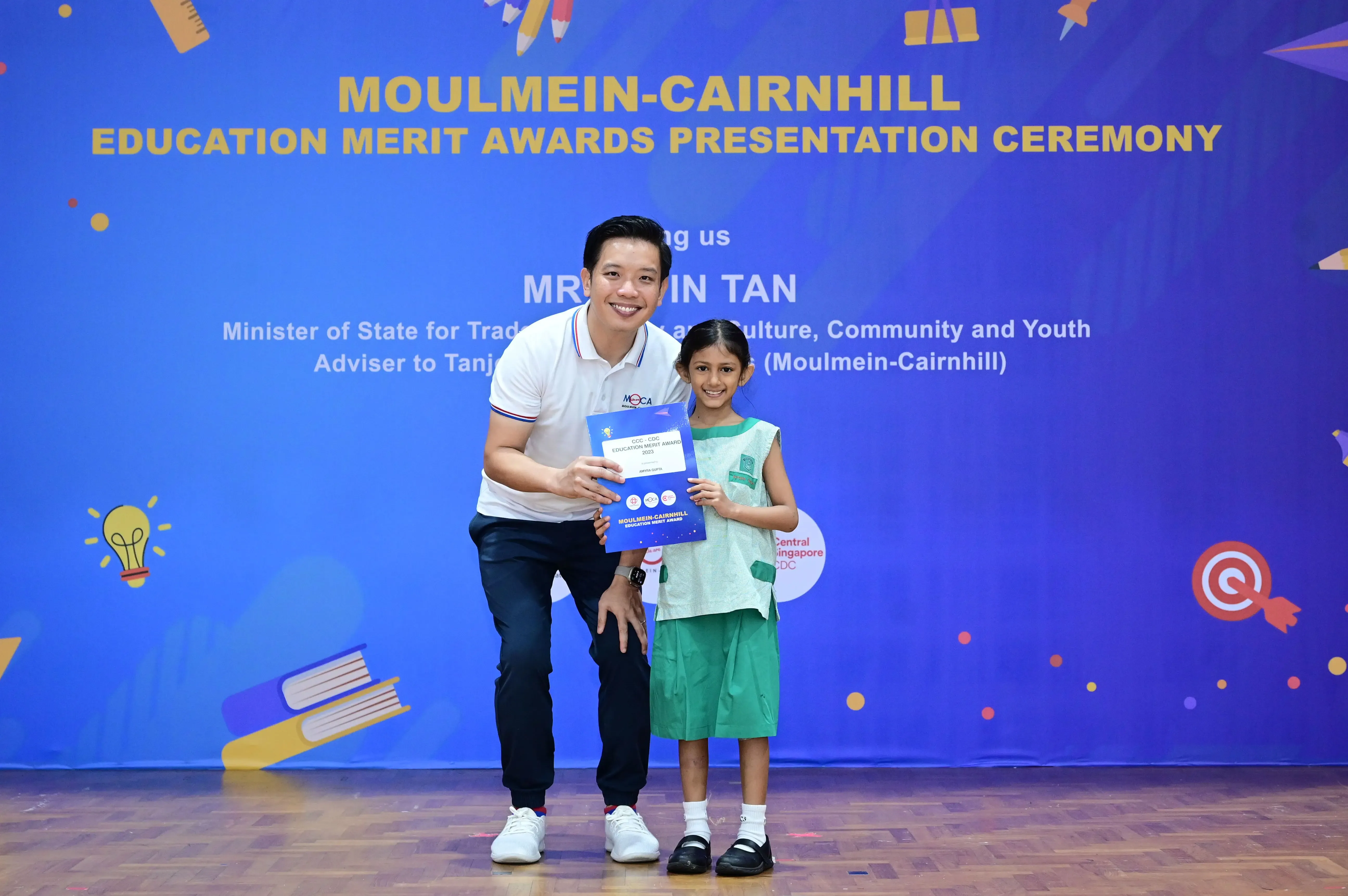 A man and a young girl hold a blue award certificate at the Moulmein-Cairnhill Education Merit Awards ceremony.