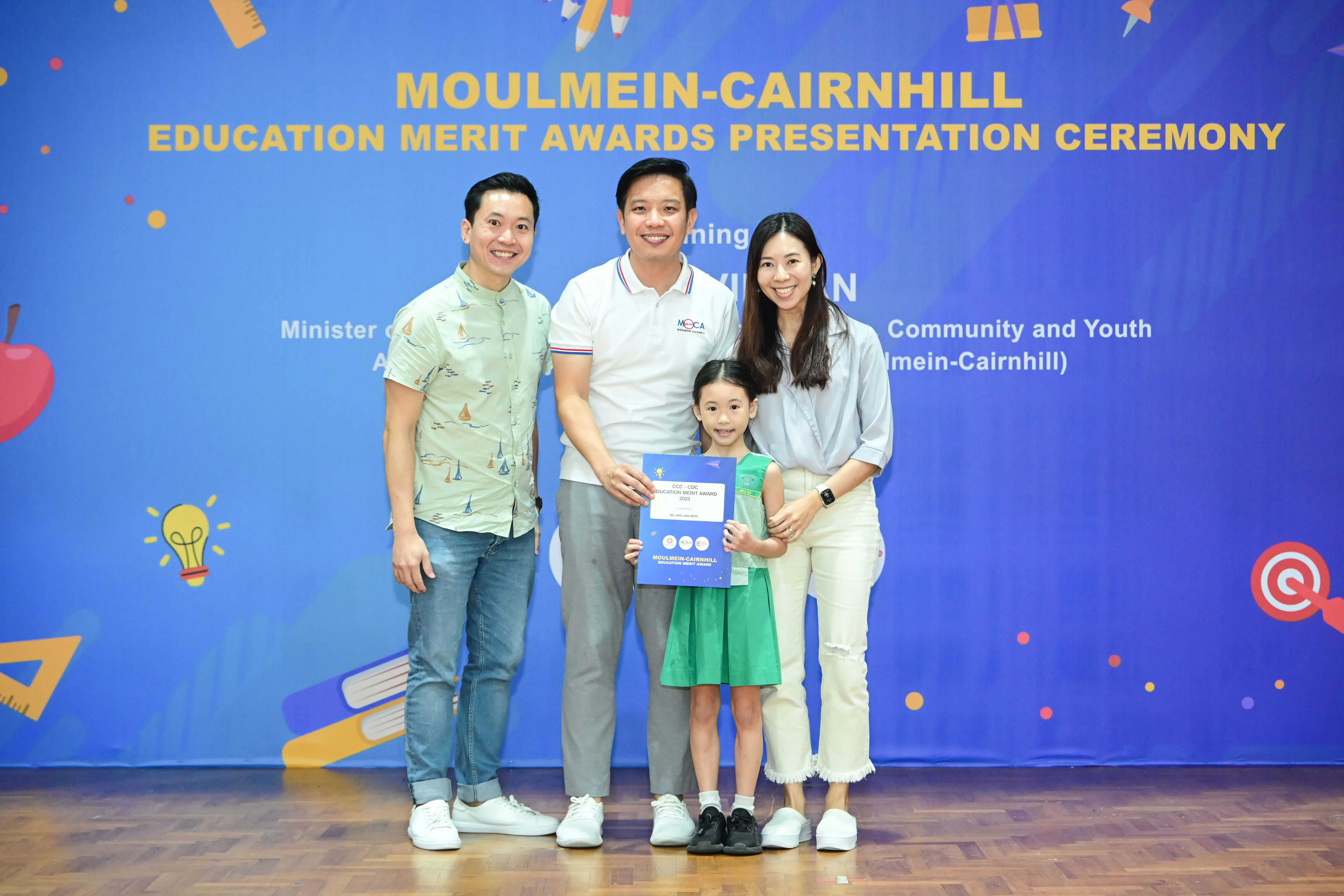 Three adults and a young girl holding a certificate pose before a Moulmein-Cairnhill Education Merit Awards banner.