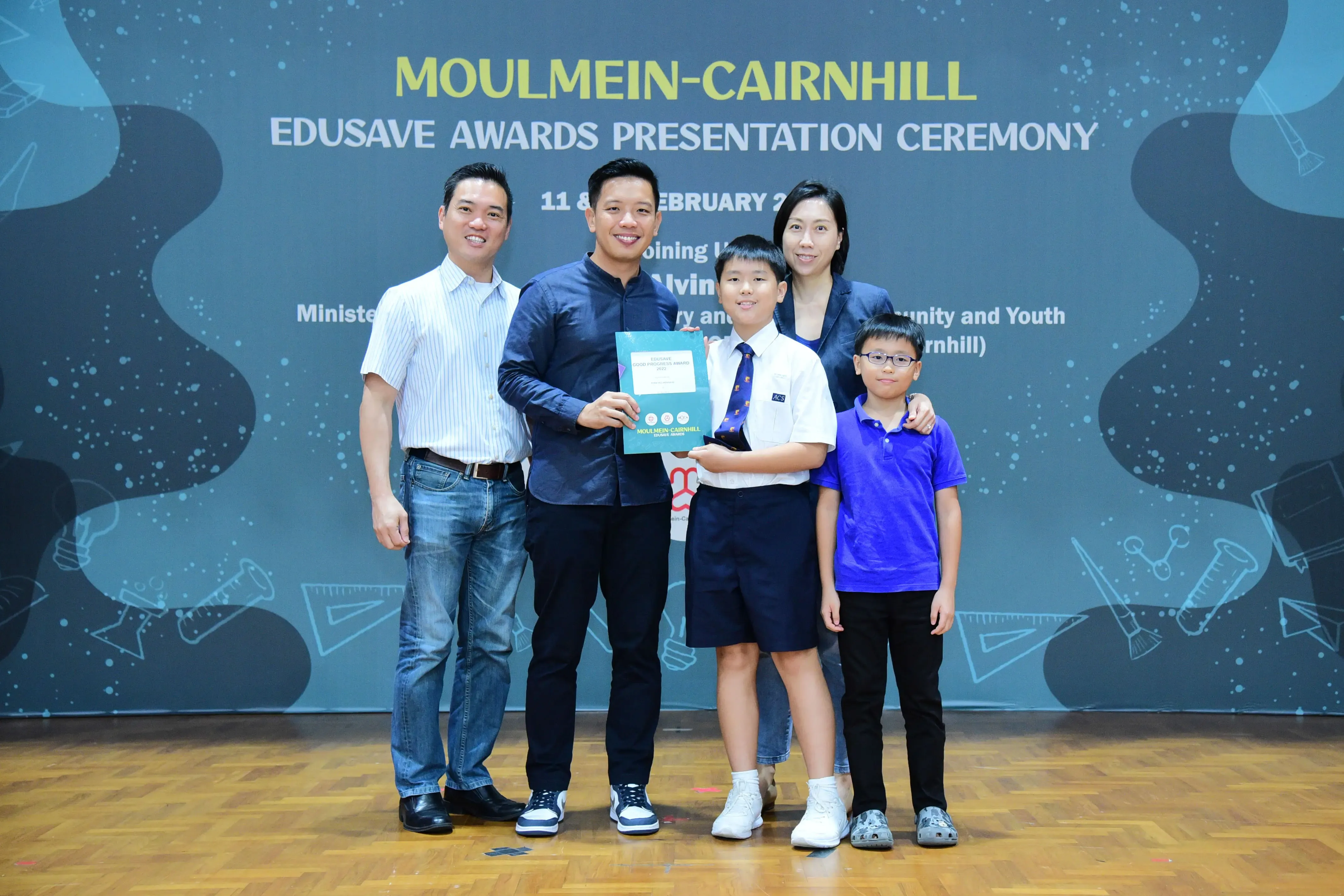Five people pose on stage at the Moulmein-Cairnhill Edusave Awards Presentation Ceremony, with a boy holding a certificate.