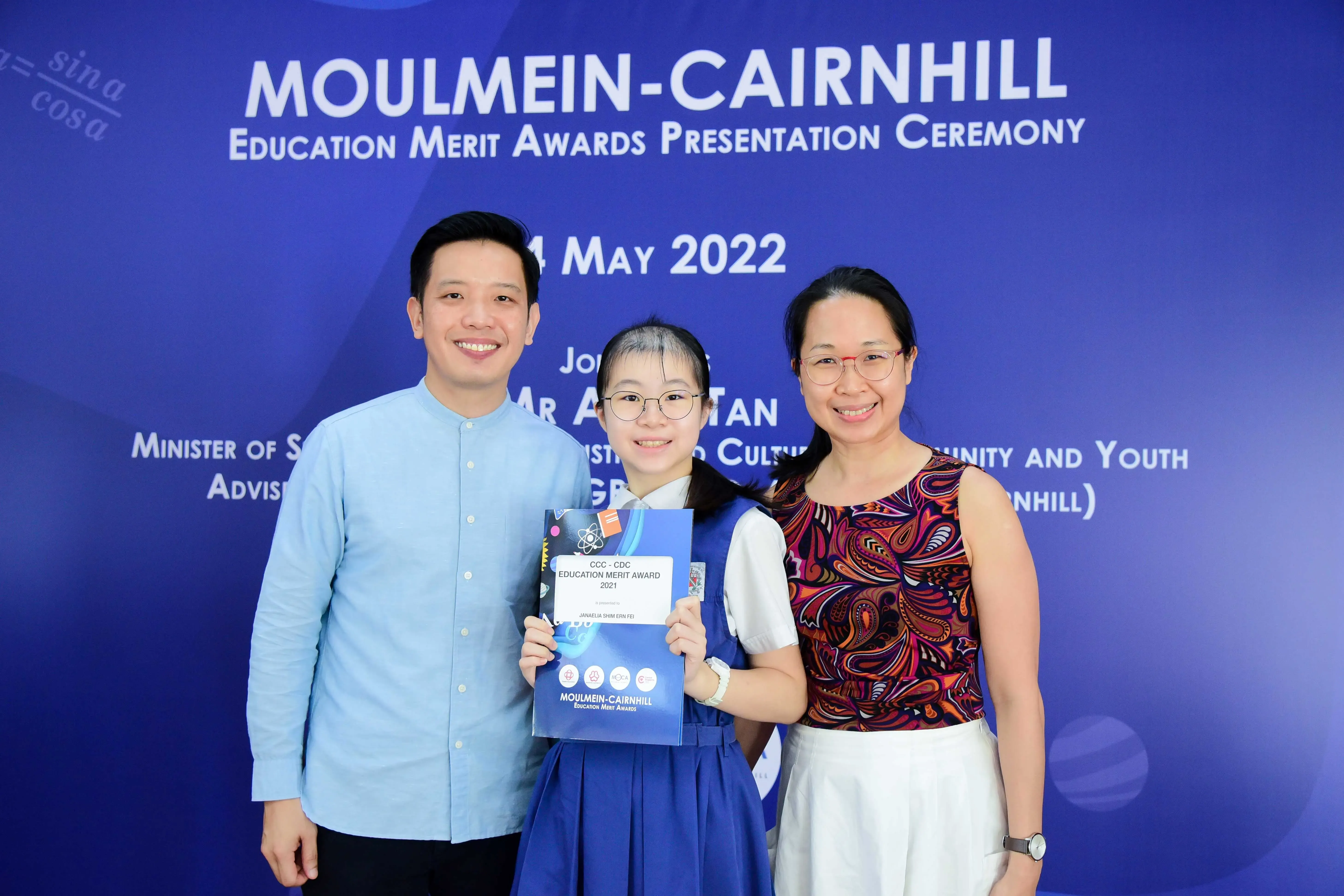 Three people smile in front of a blue Moulmein-Cairnhill Education Merit Awards banner dated May 2022.