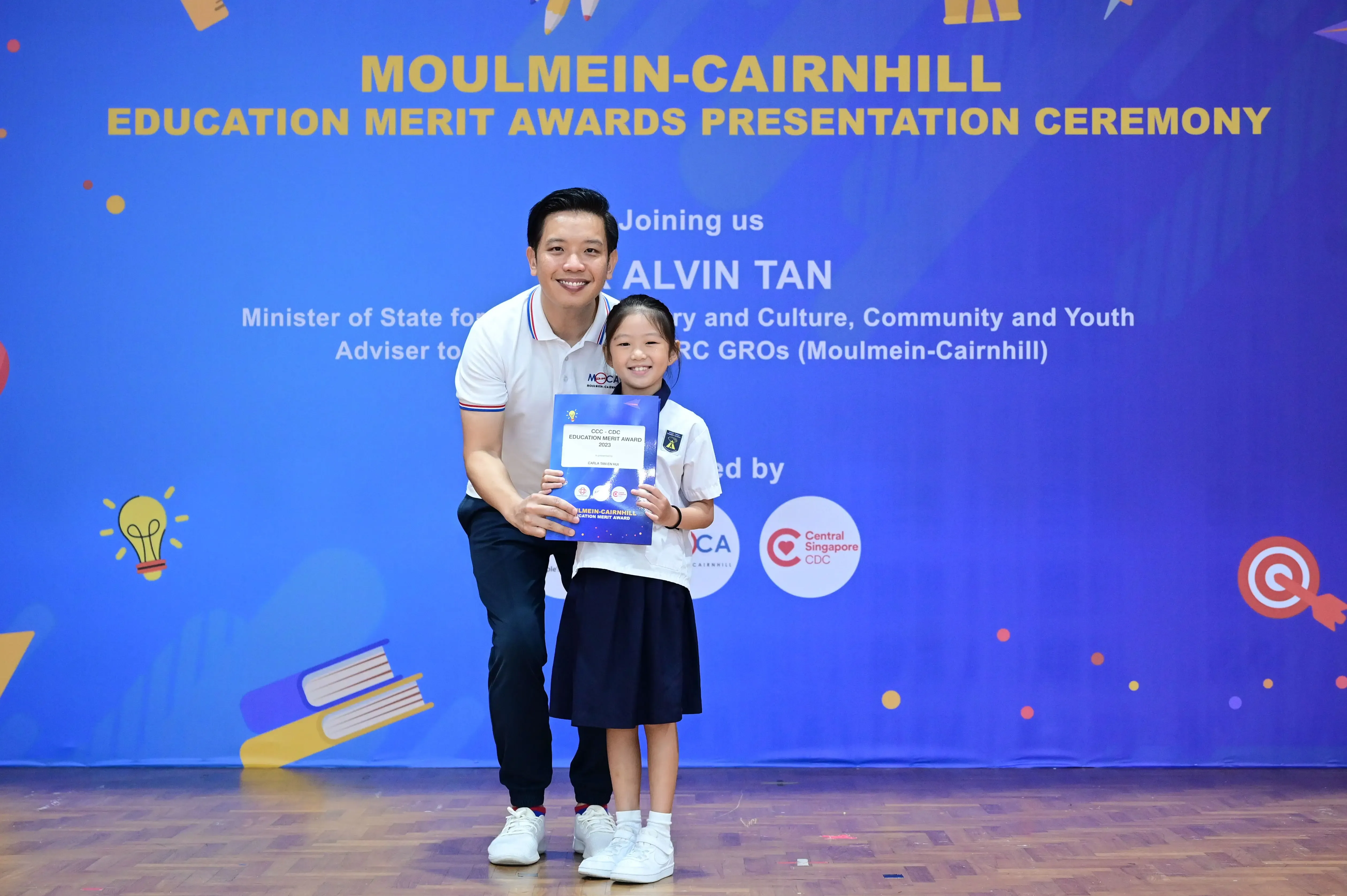 A man and a young girl hold a certificate together in front of a Moulmein-Cairnhill Education Merit Awards banner.