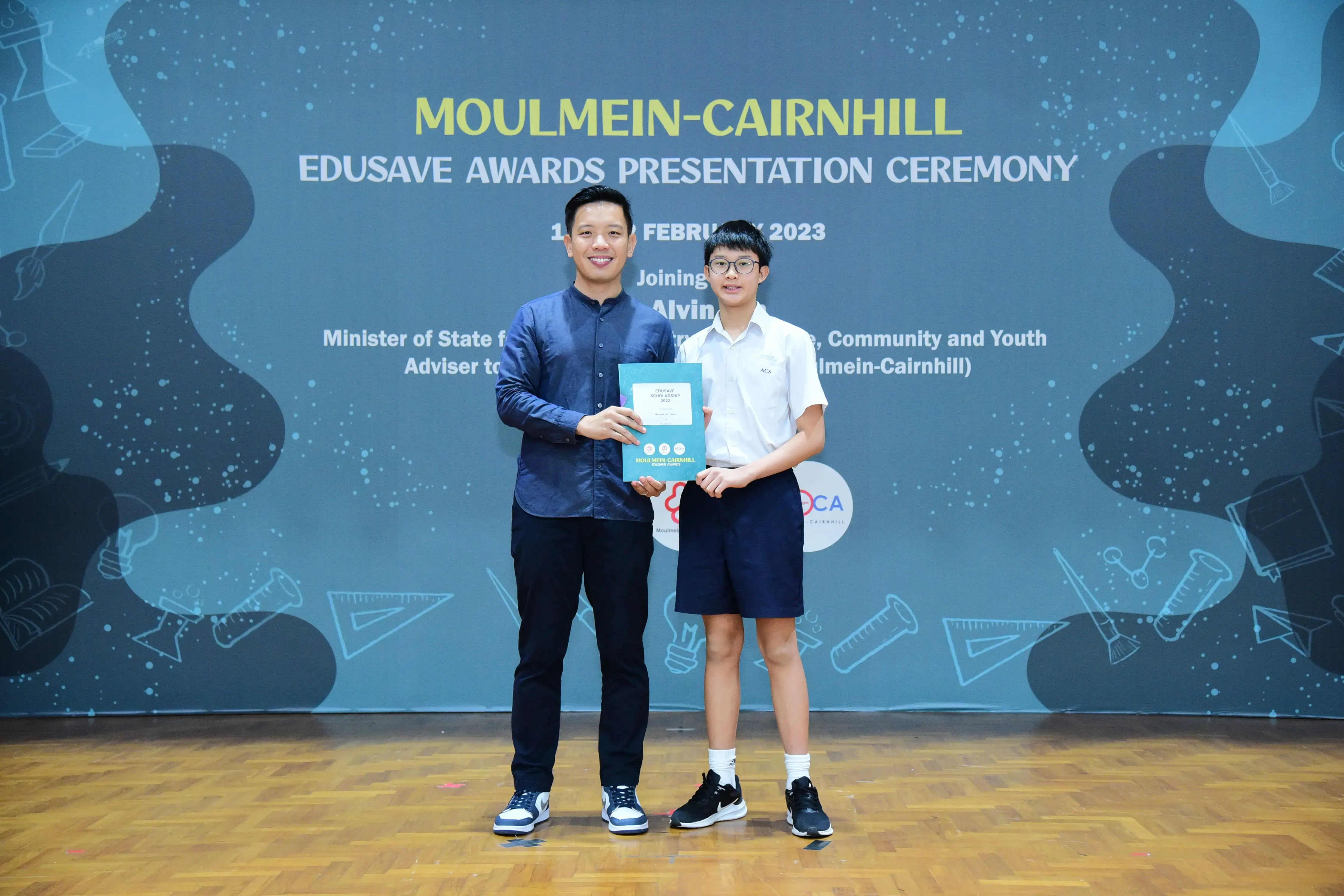 Two people hold a certificate together on stage at the Moulmein-Cairnhill Edusave Awards Presentation Ceremony 2023.