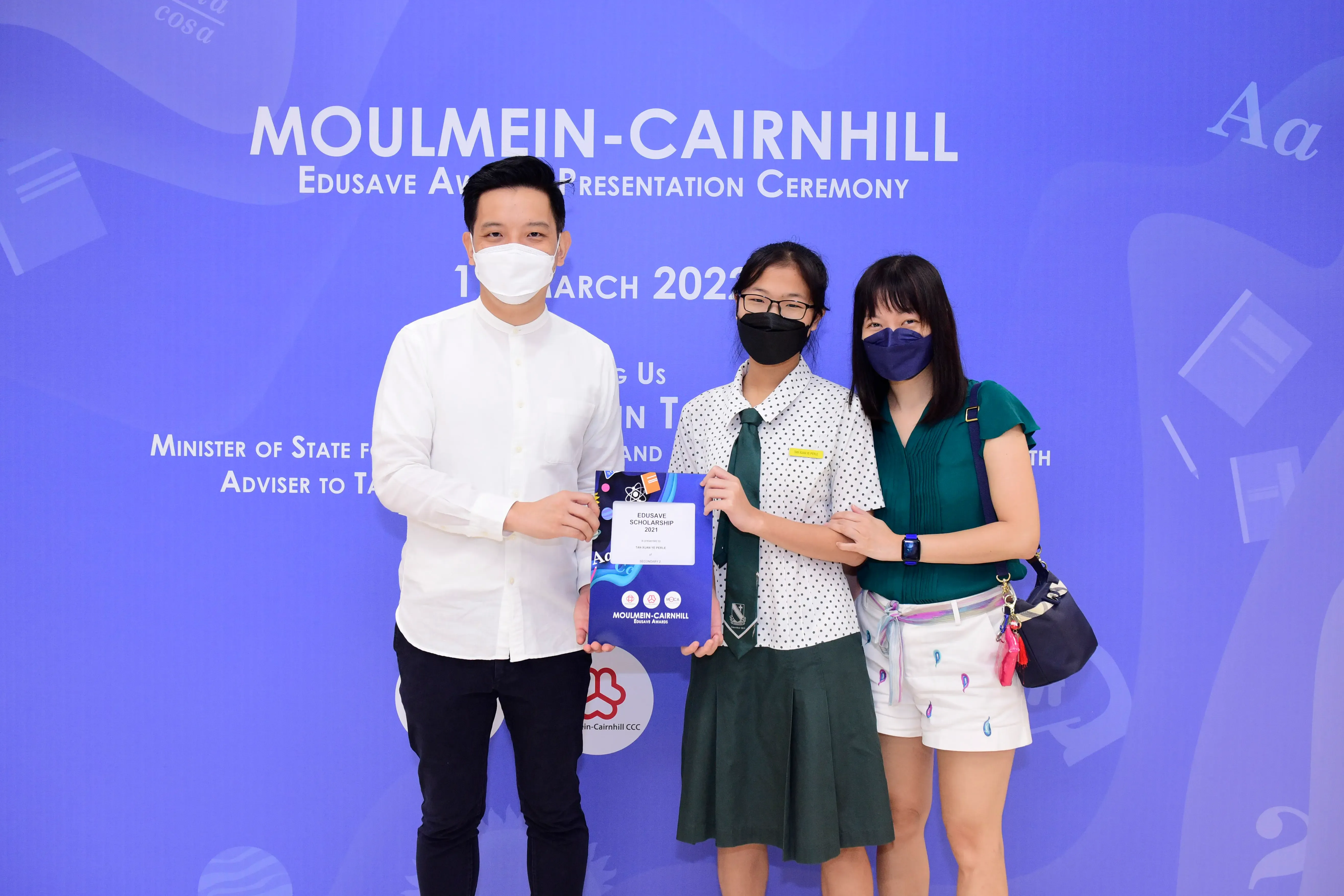Three masked people pose at the Moulmein-Cairnhill Edusave Award Presentation Ceremony, center person holds a certificate.