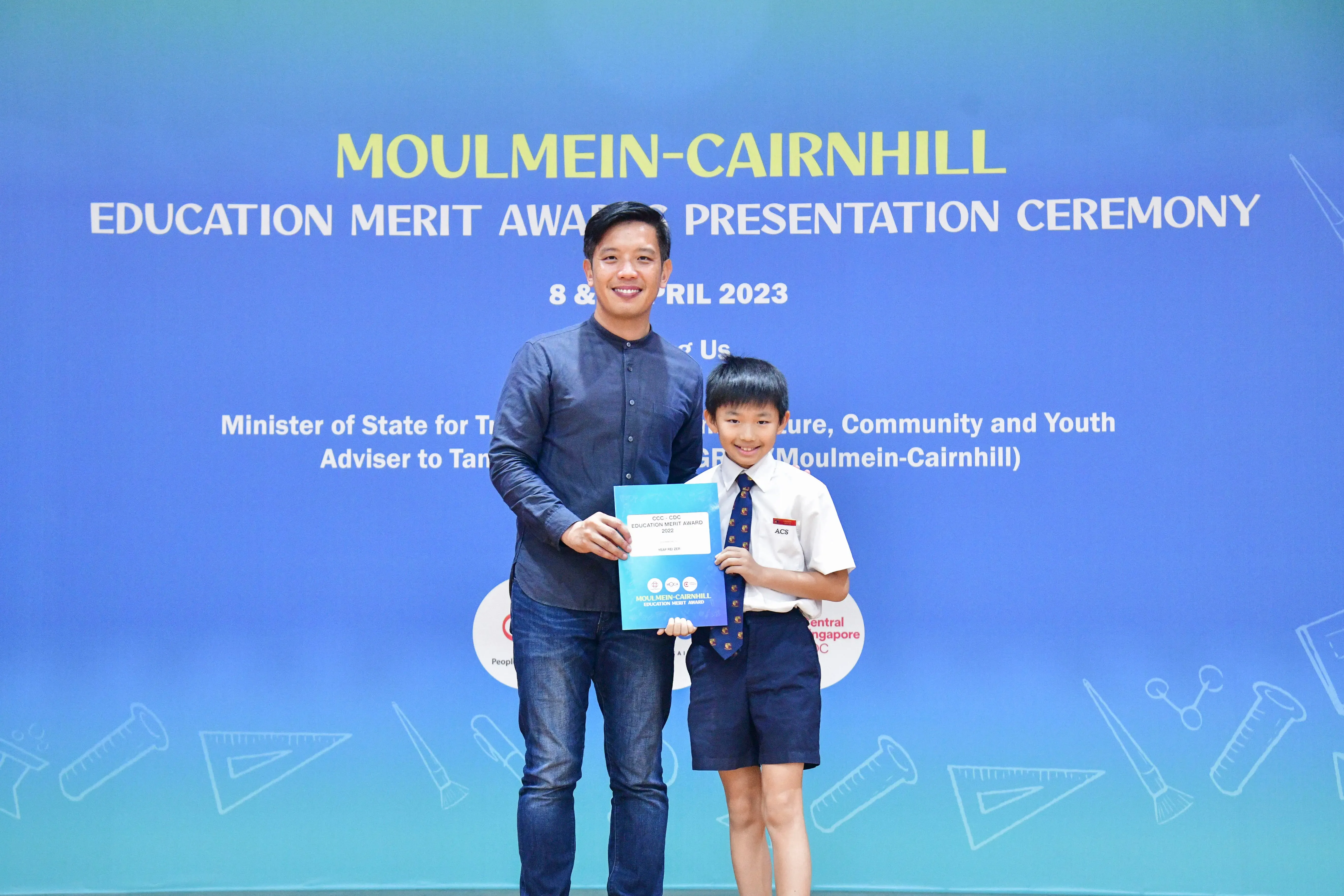 A man and a young boy in school uniform hold a certificate at the Moulmein-Cairnhill Education Merit Award ceremony.