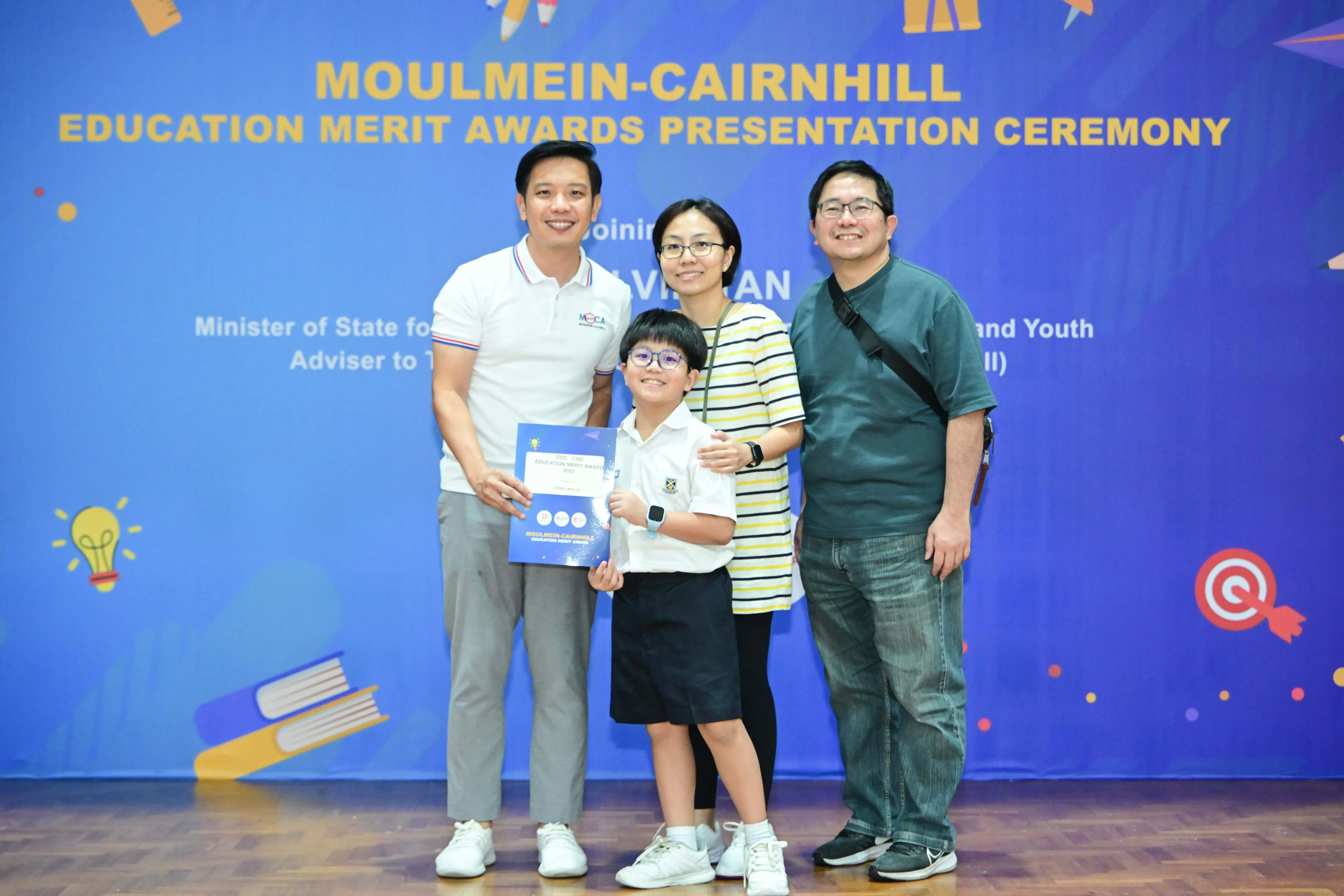 Three adults and a child pose at the Moulmein-Cairnhill Education Merit Awards, with the child holding a certificate.