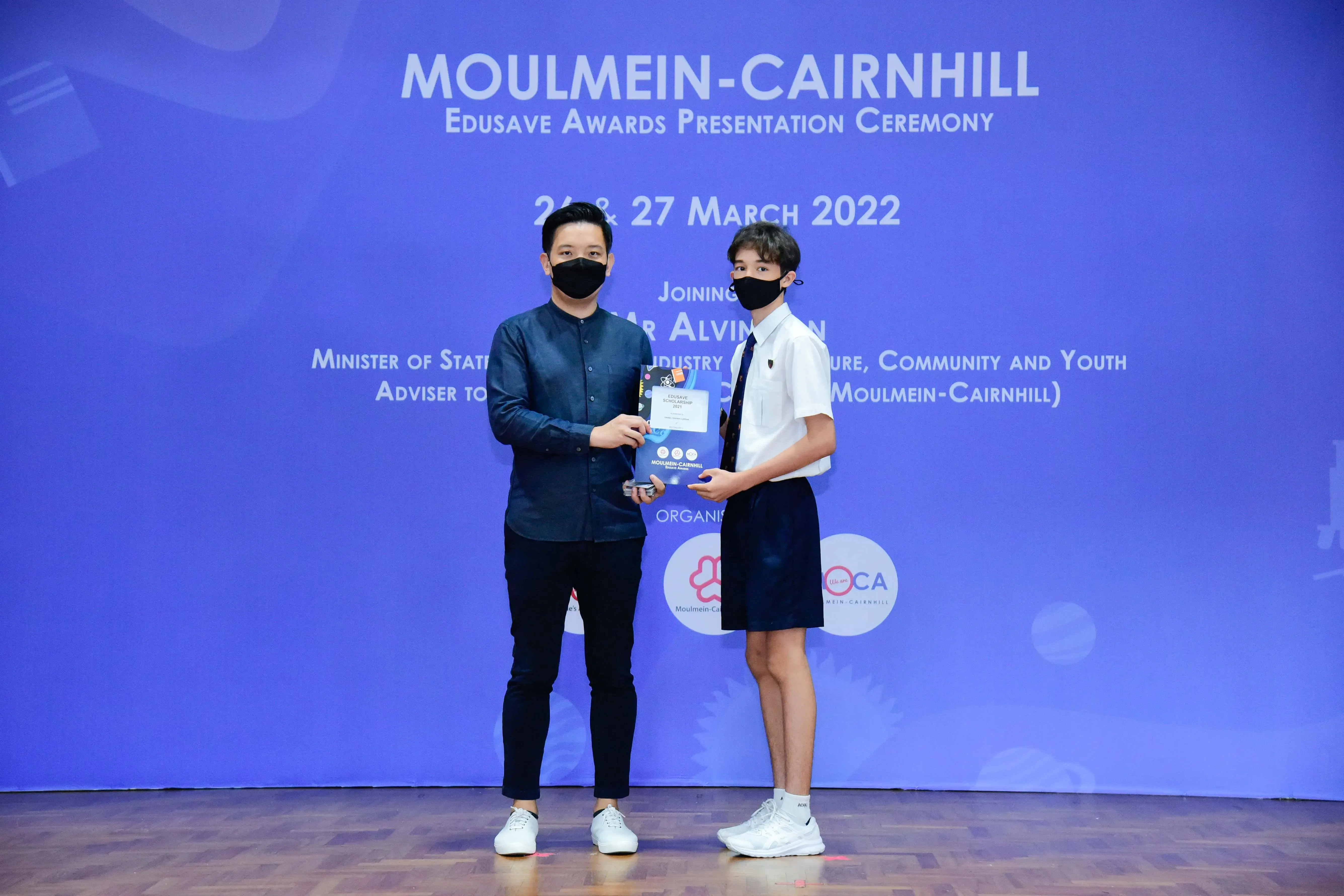 Two masked individuals hold an award at the Moulmein-Cairnhill Edusave Awards Presentation Ceremony, March 2022.
