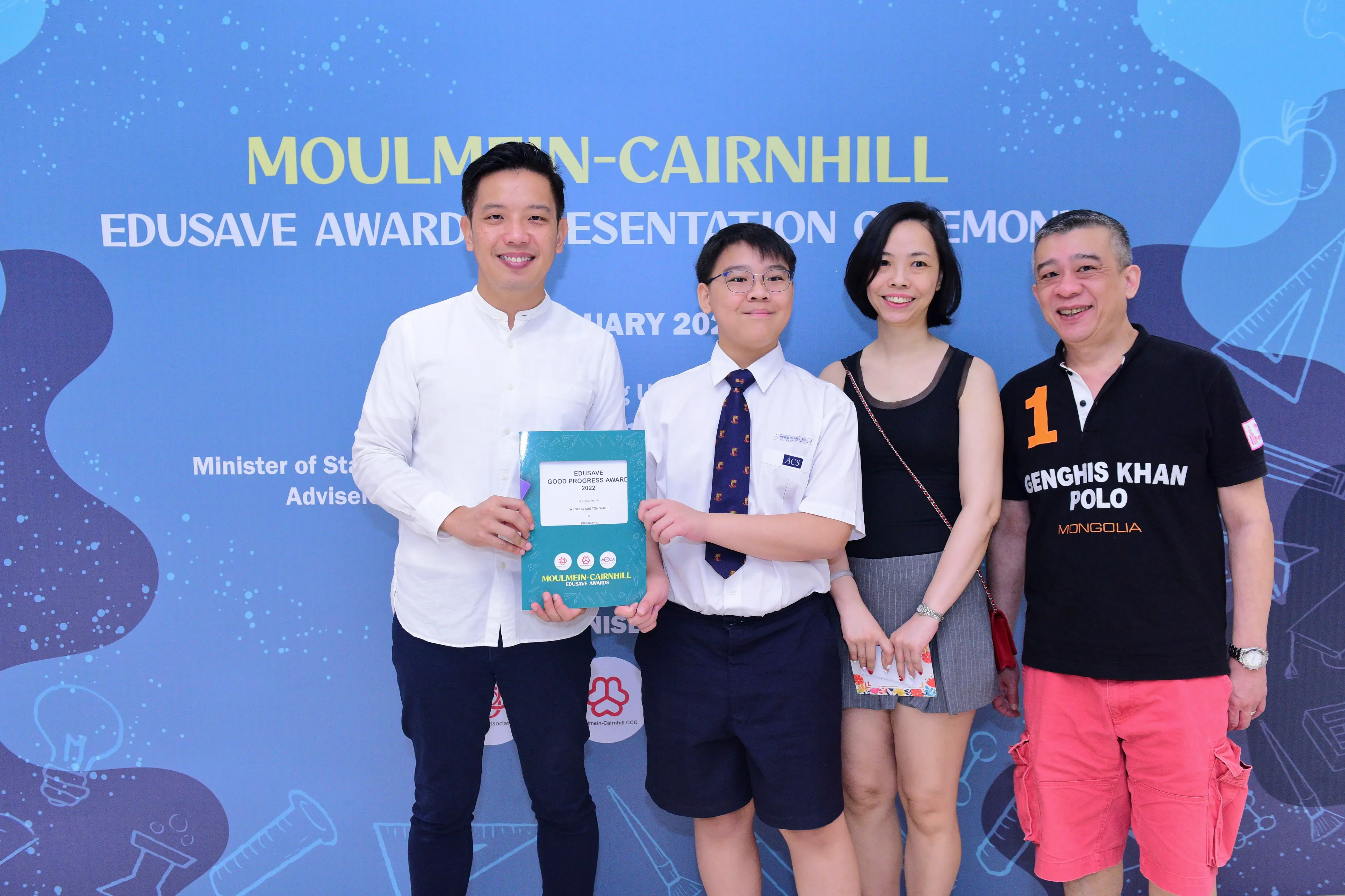 Four people smiling at the Moulmein-Cairnhill Edusave Awards ceremony, with a student holding a teal award folder.