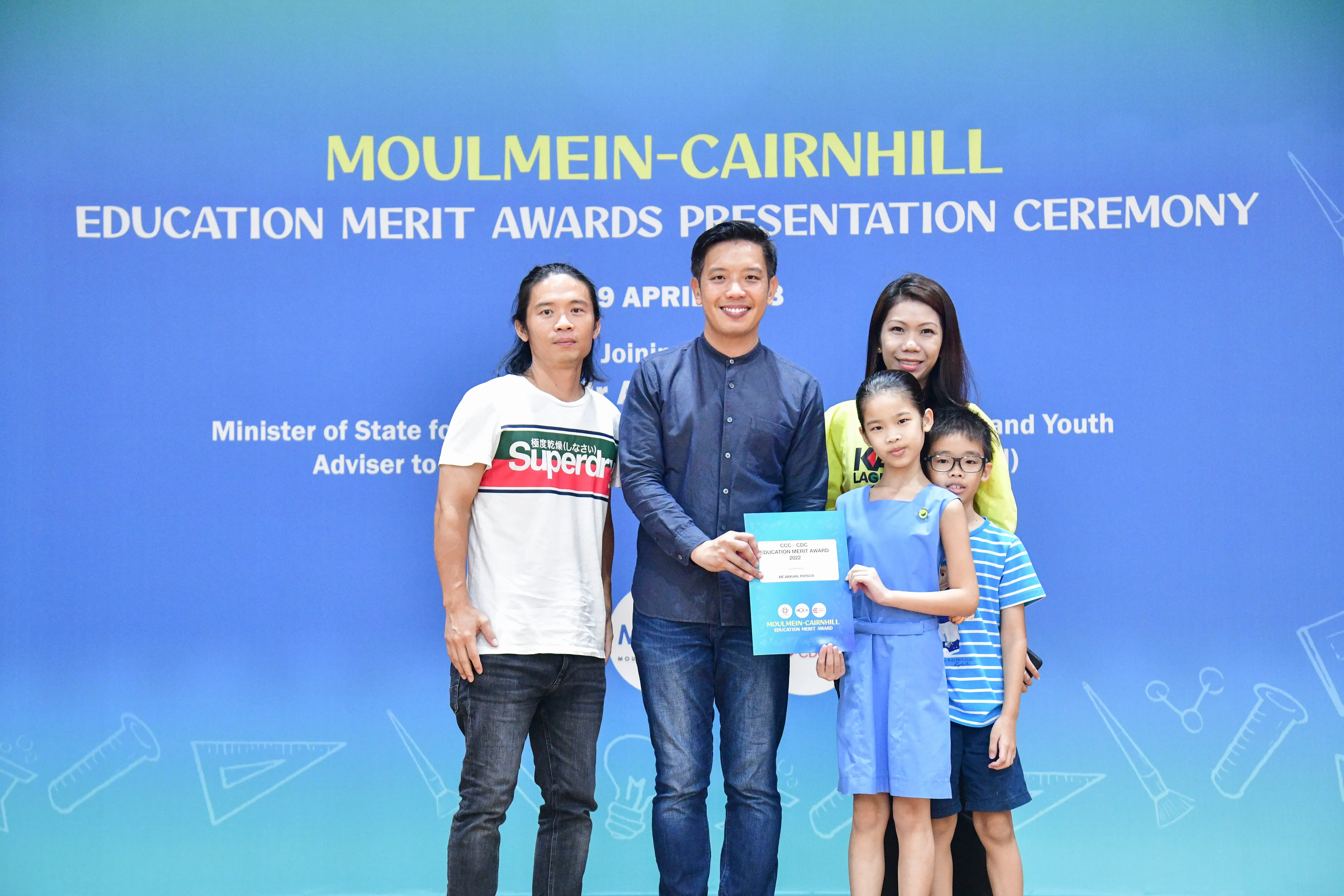 Five people pose at the Moulmein-Cairnhill Education Merit Awards Presentation Ceremony, with a girl holding a certificate.