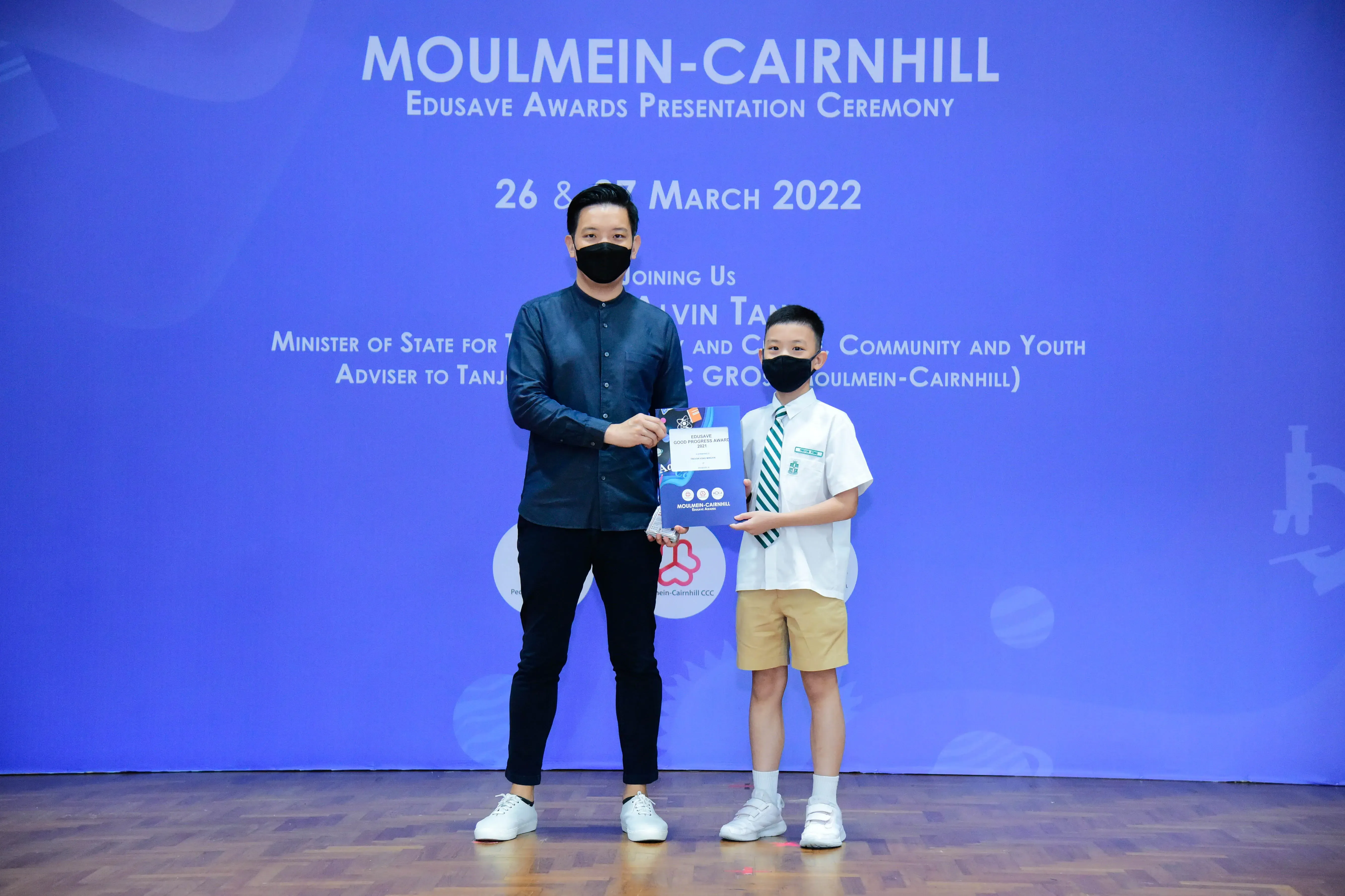 An adult and a young student in uniform hold a certificate at the Moulmein-Cairnhill Edusave Awards Ceremony, March 2022.