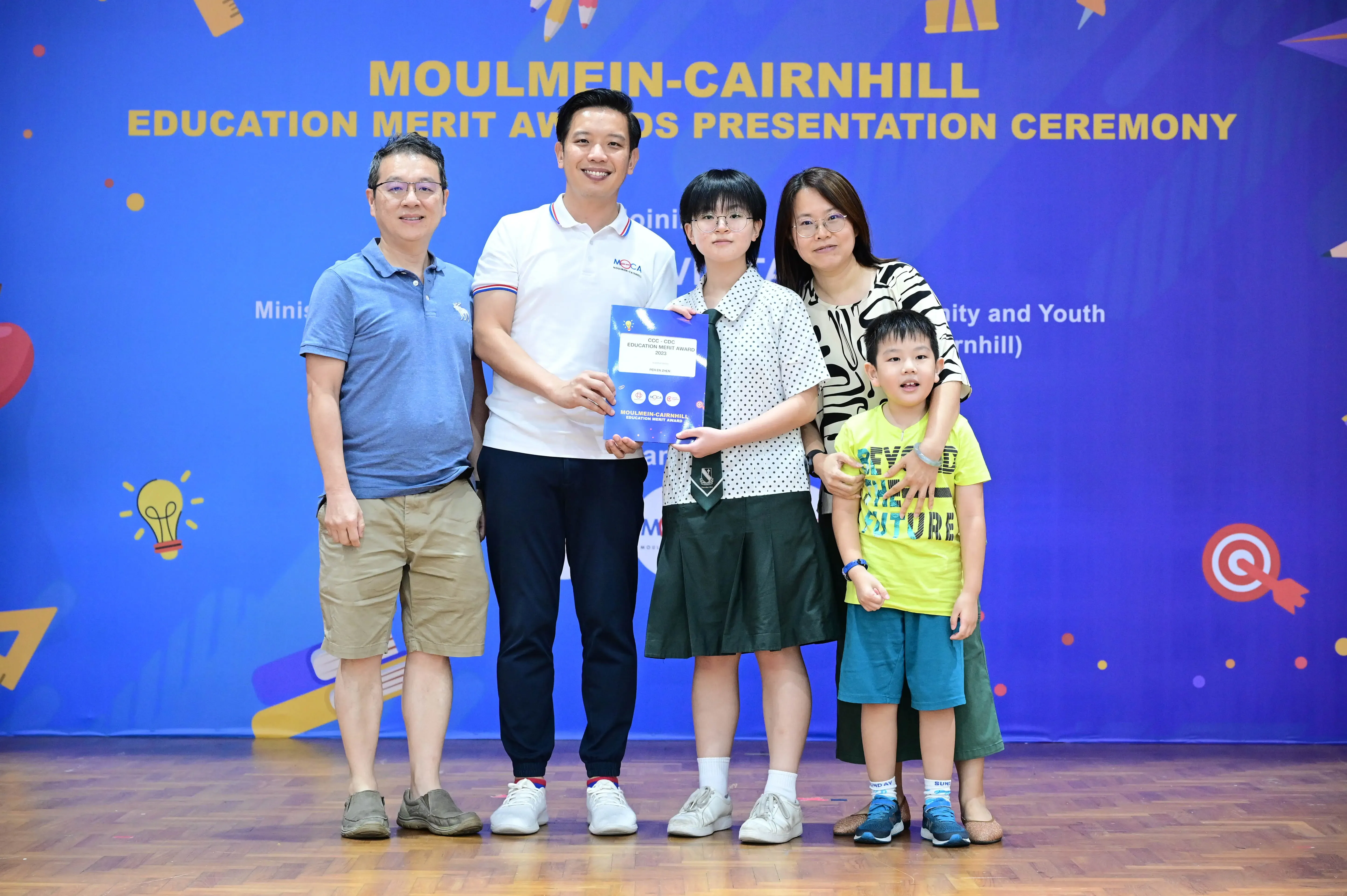 Five people pose at the Moulmein-Cairnhill Education Merit Awards Presentation Ceremony, with a girl holding an award certificate.