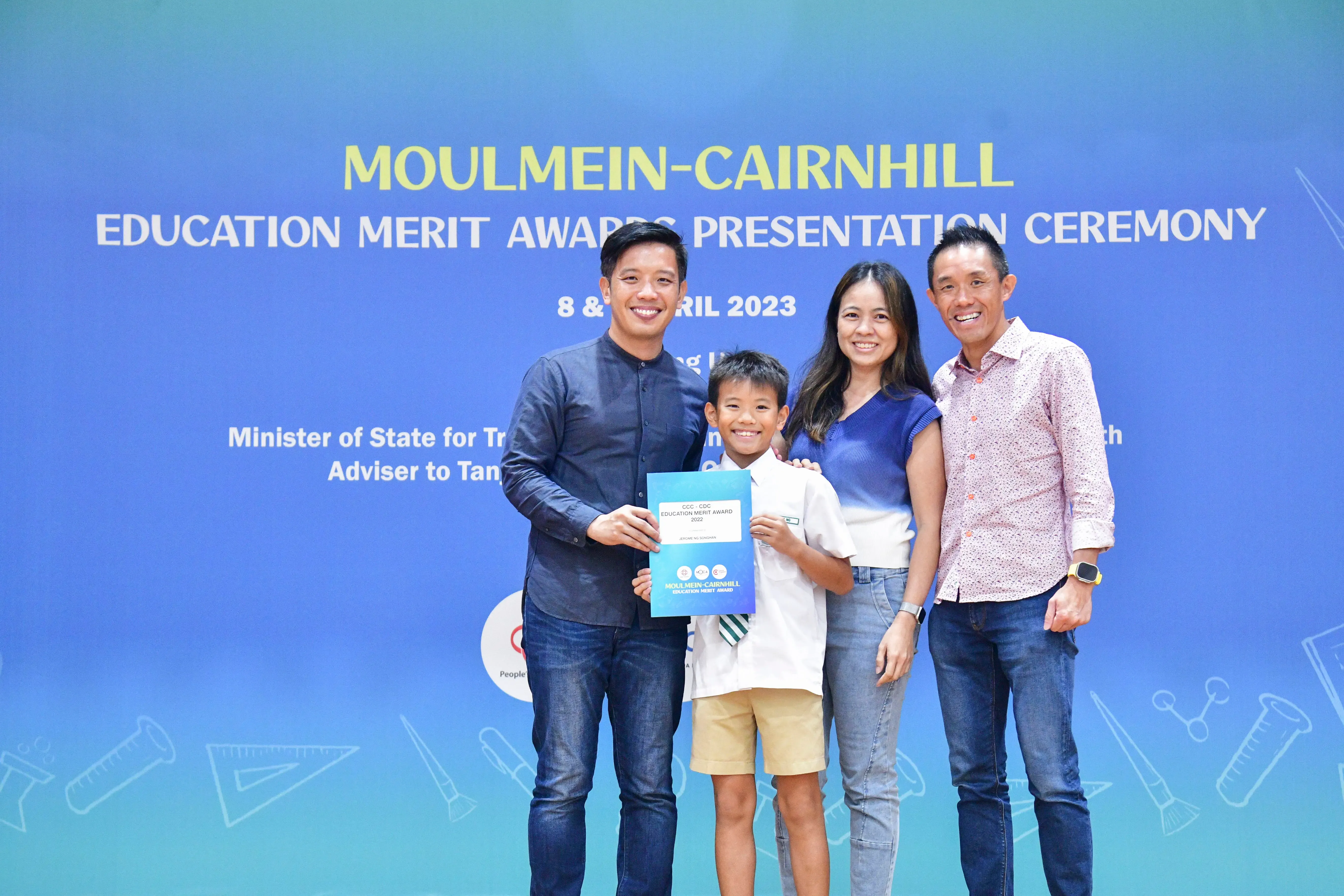 A smiling boy holds a certificate with three adults at the Moulmein-Cairnhill Education Merit Awards Ceremony 2023.