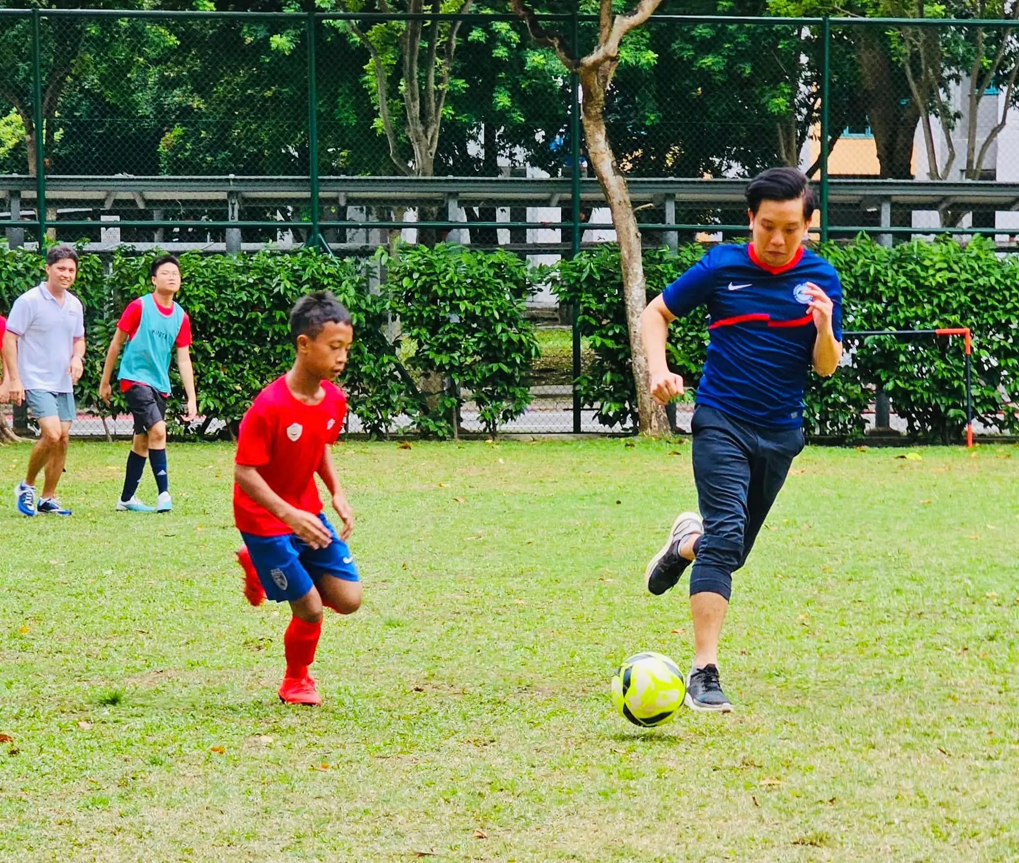 A man in a blue jersey kicks a soccer ball while a boy in red defends on a grass field.