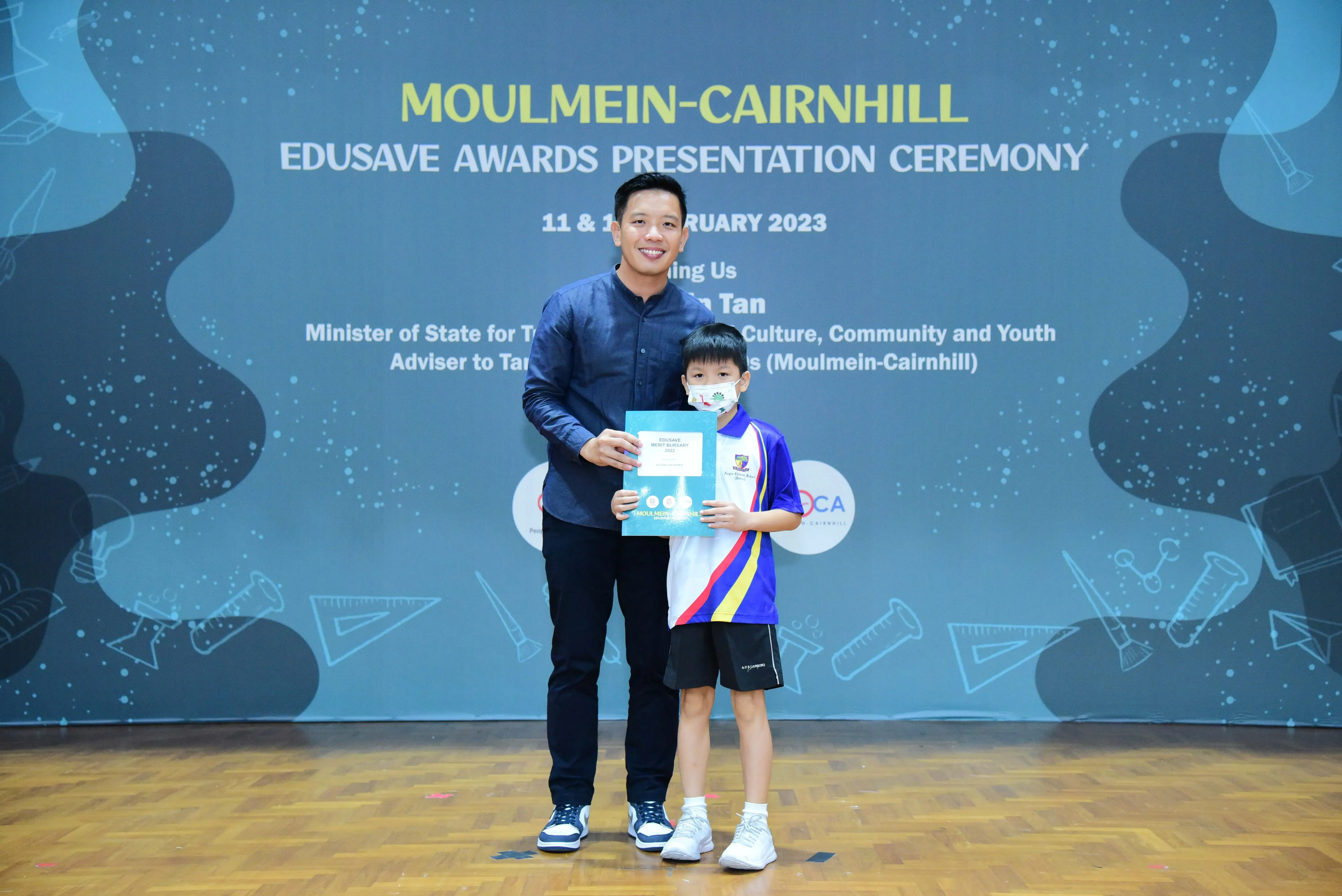 A man and a masked young boy hold a certificate at the Moulmein-Cairnhill Edusave Awards Ceremony 2023.