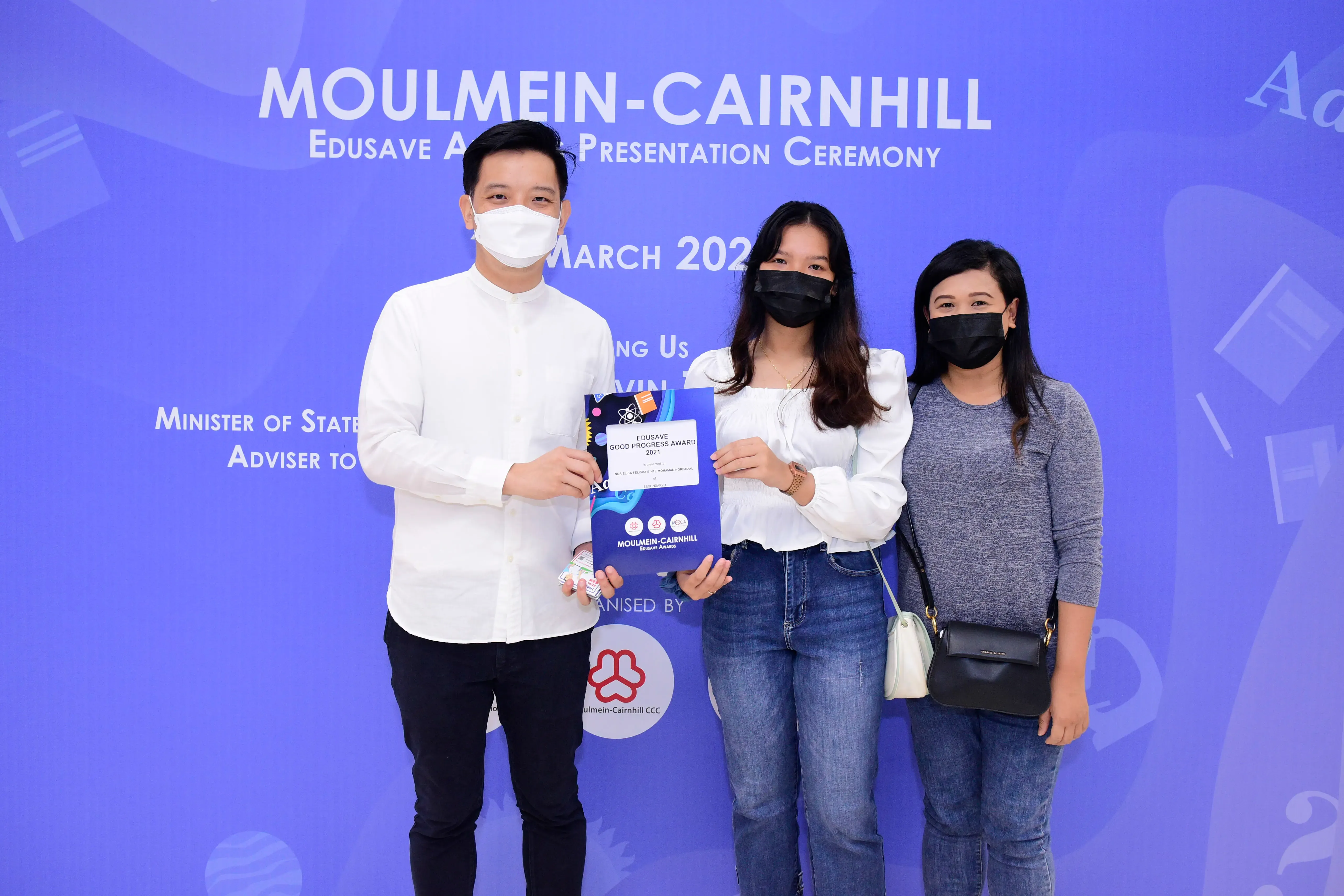 Three masked people pose at the Moulmein-Cairnhill Edusave Award Presentation Ceremony, holding an award certificate.