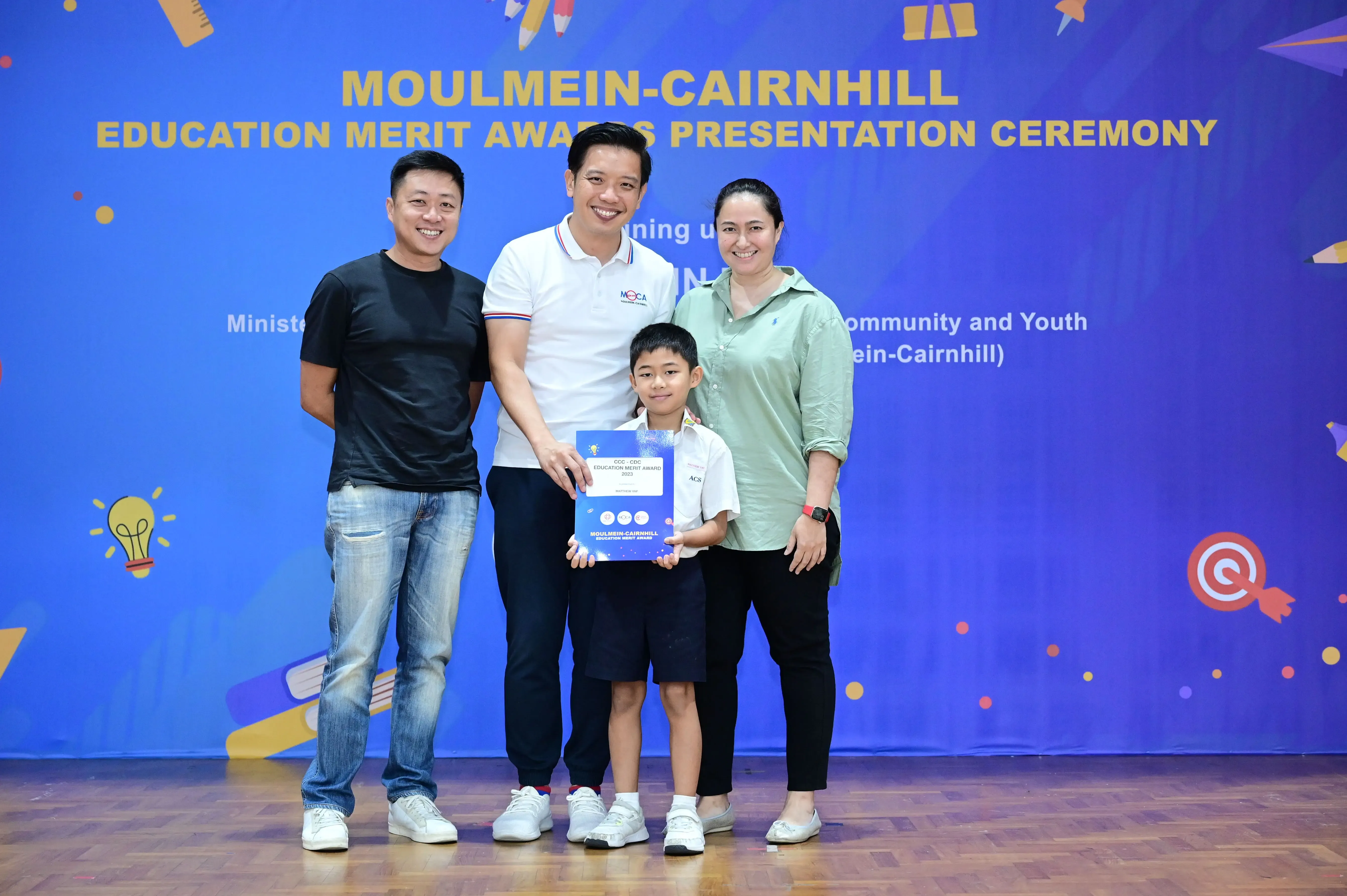 Three adults and a child pose at the Moulmein-Cairnhill Education Merit Awards ceremony, child holding a certificate.
