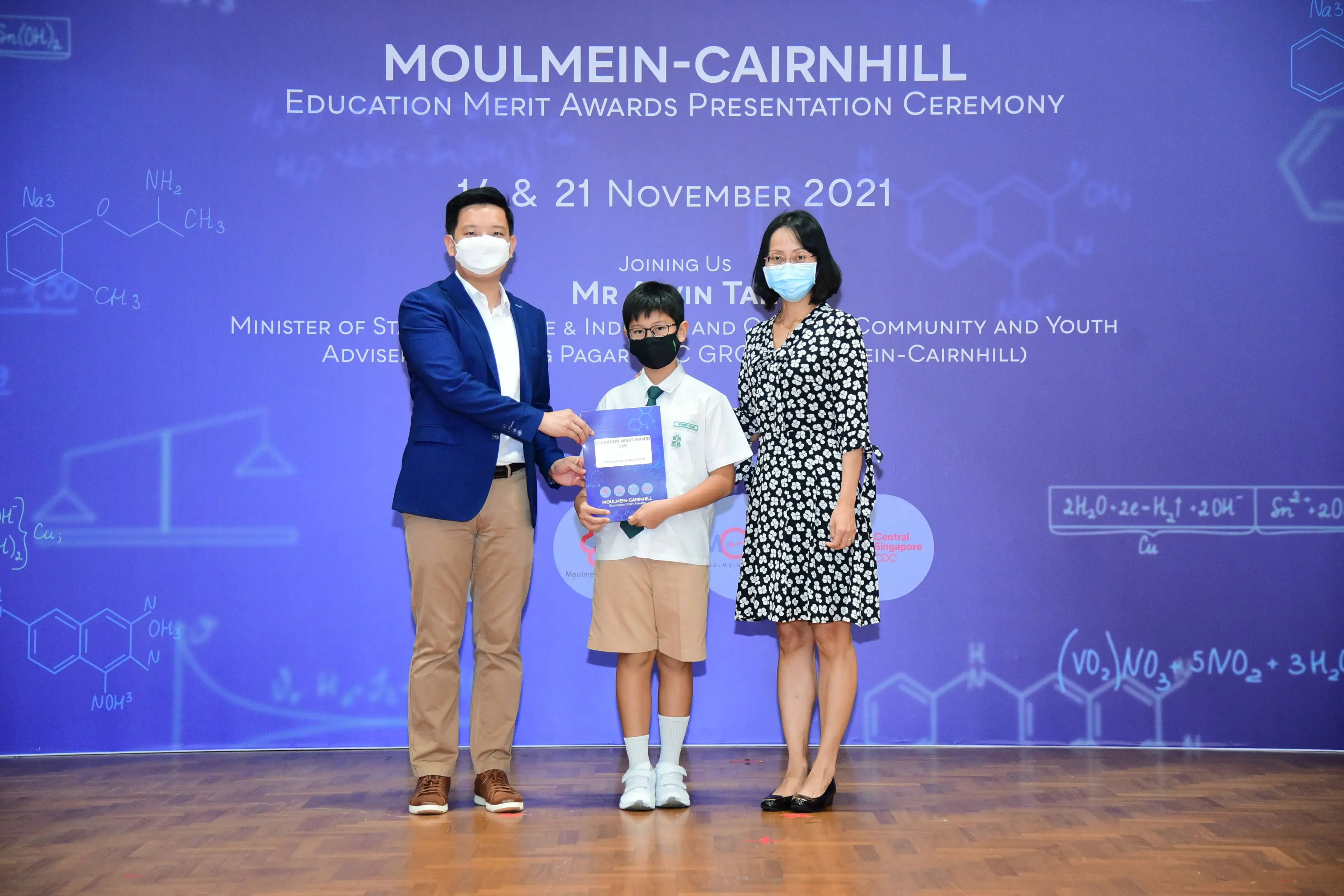 Two adults and a young student in school uniform hold an award at the Moulmein-Cairnhill Education Merit Awards 2021.