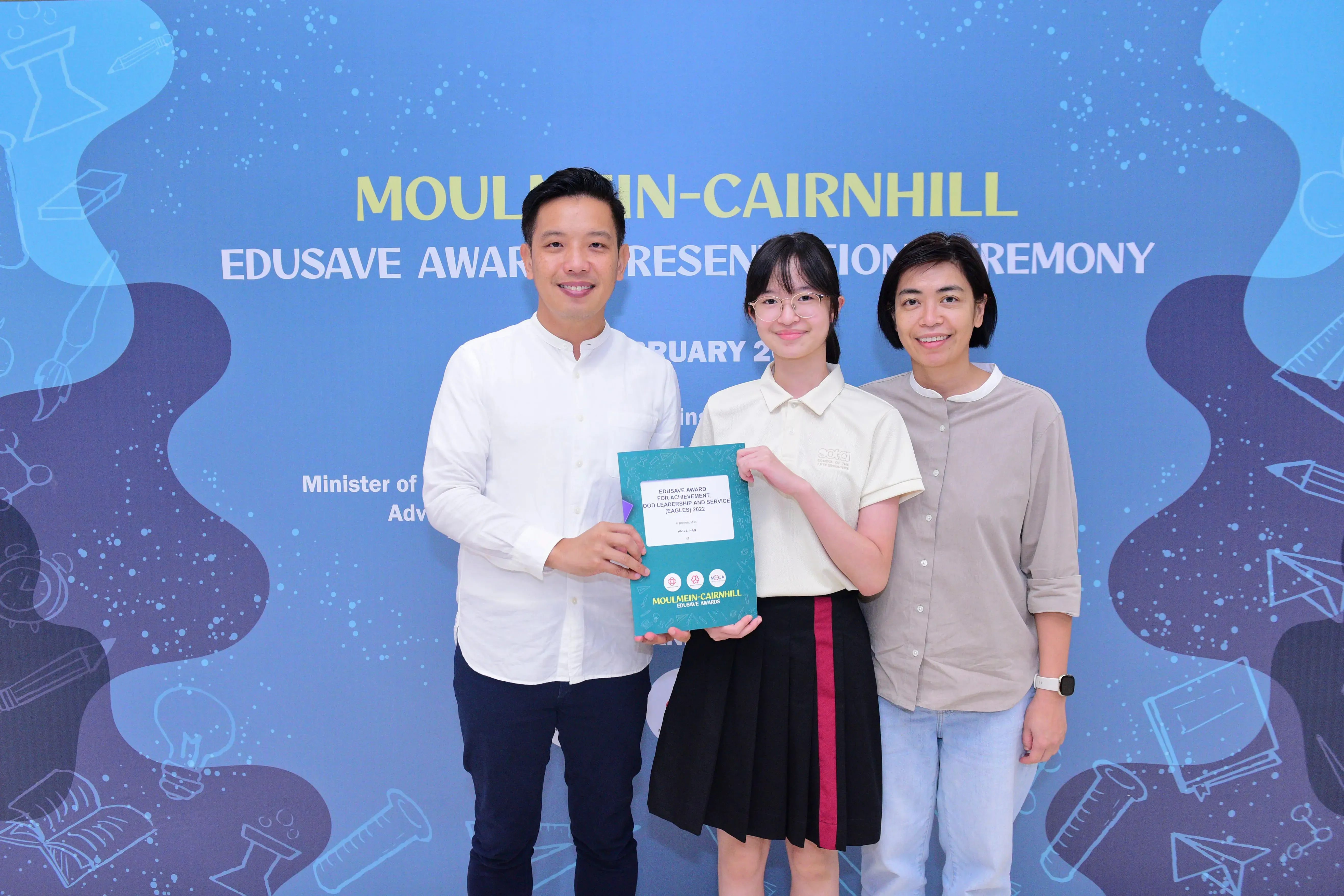Three people smile while holding a certificate at the Moulmein-Cairnhill Edusave Award Presentation Ceremony.
