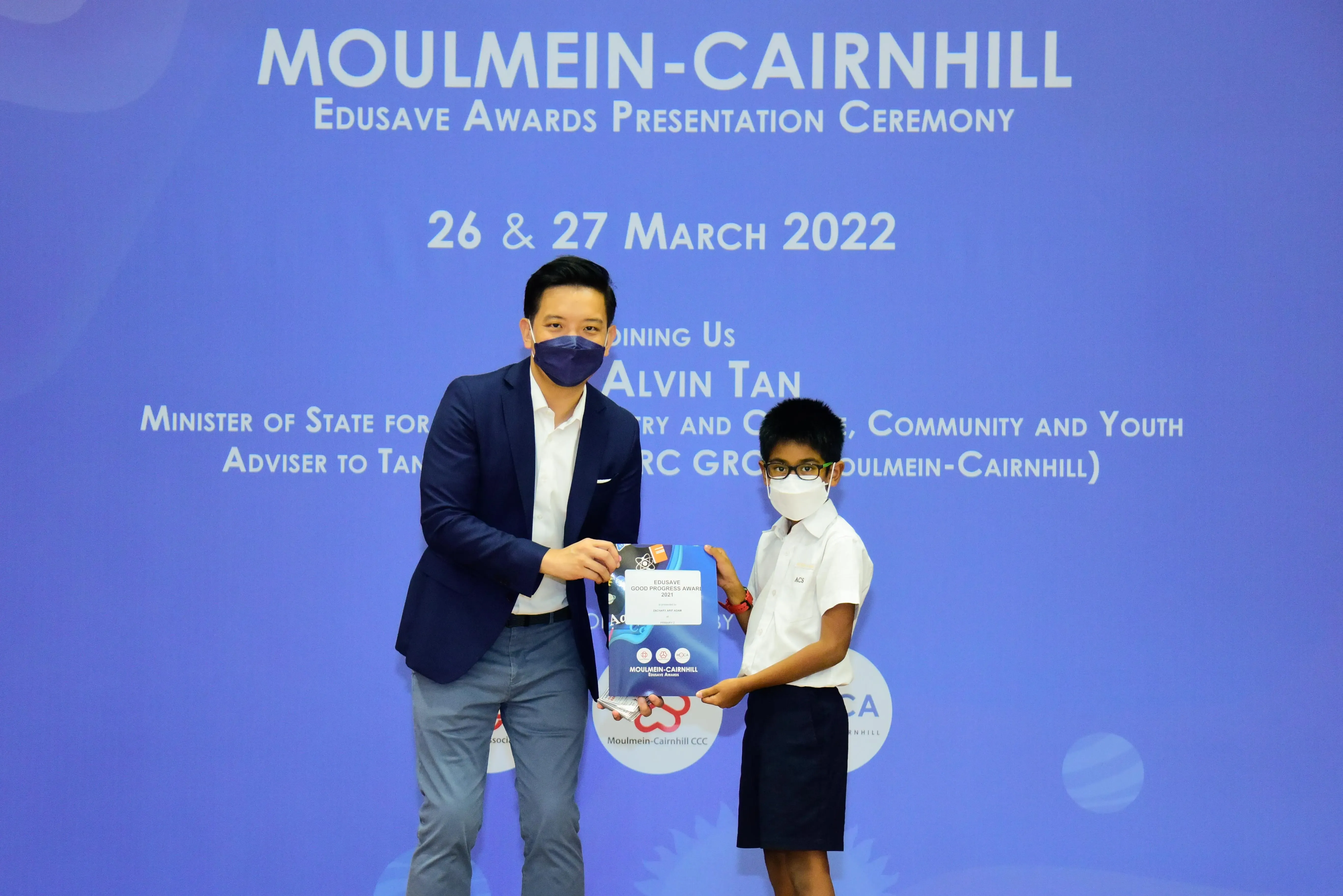 An adult in a suit presents an award to a masked child at the Moulmein-Cairnhill Edusave Awards Ceremony, March 2022.