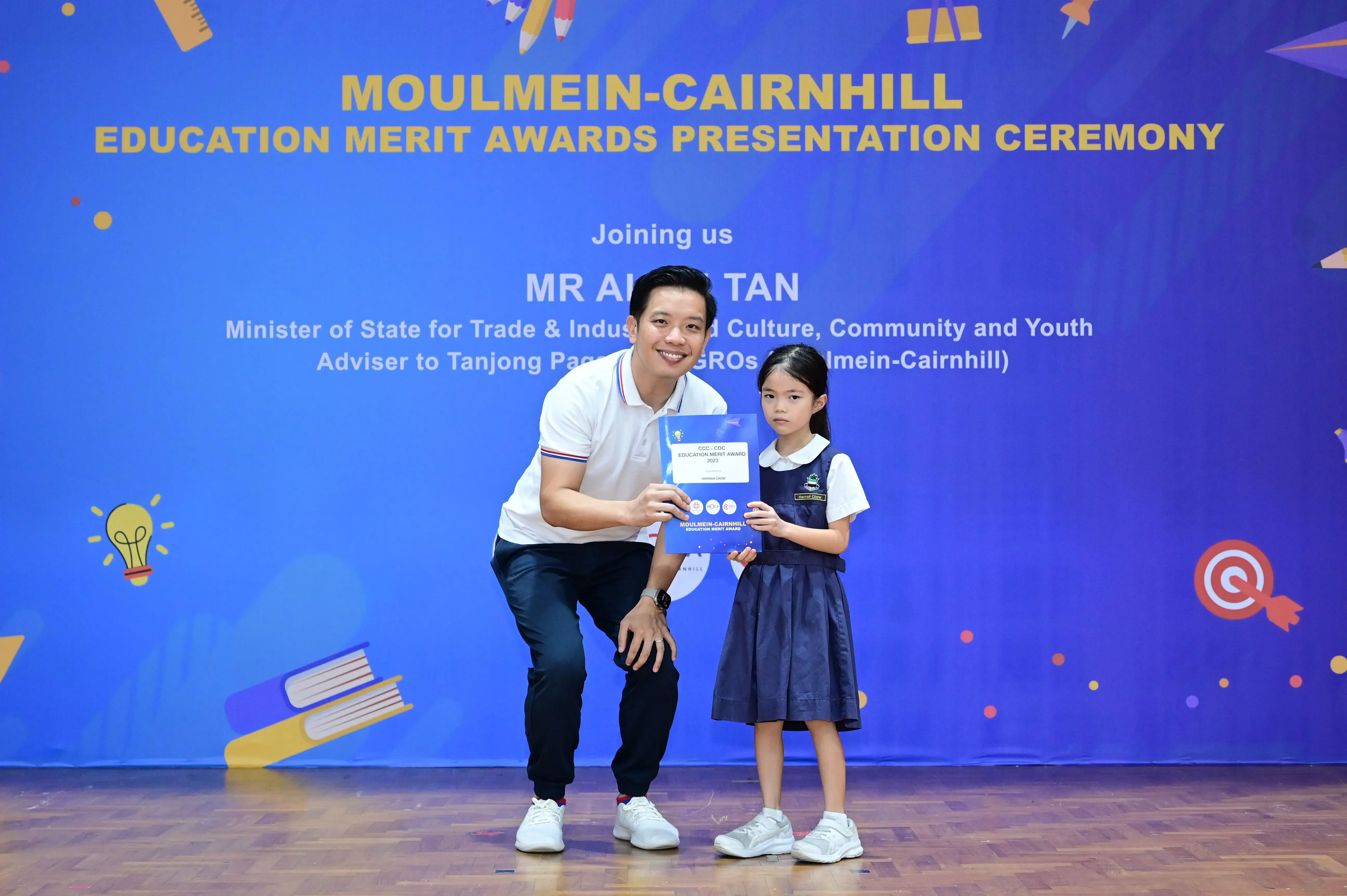 A man kneeling beside a young girl in school uniform, both holding an award certificate at an education merit ceremony.