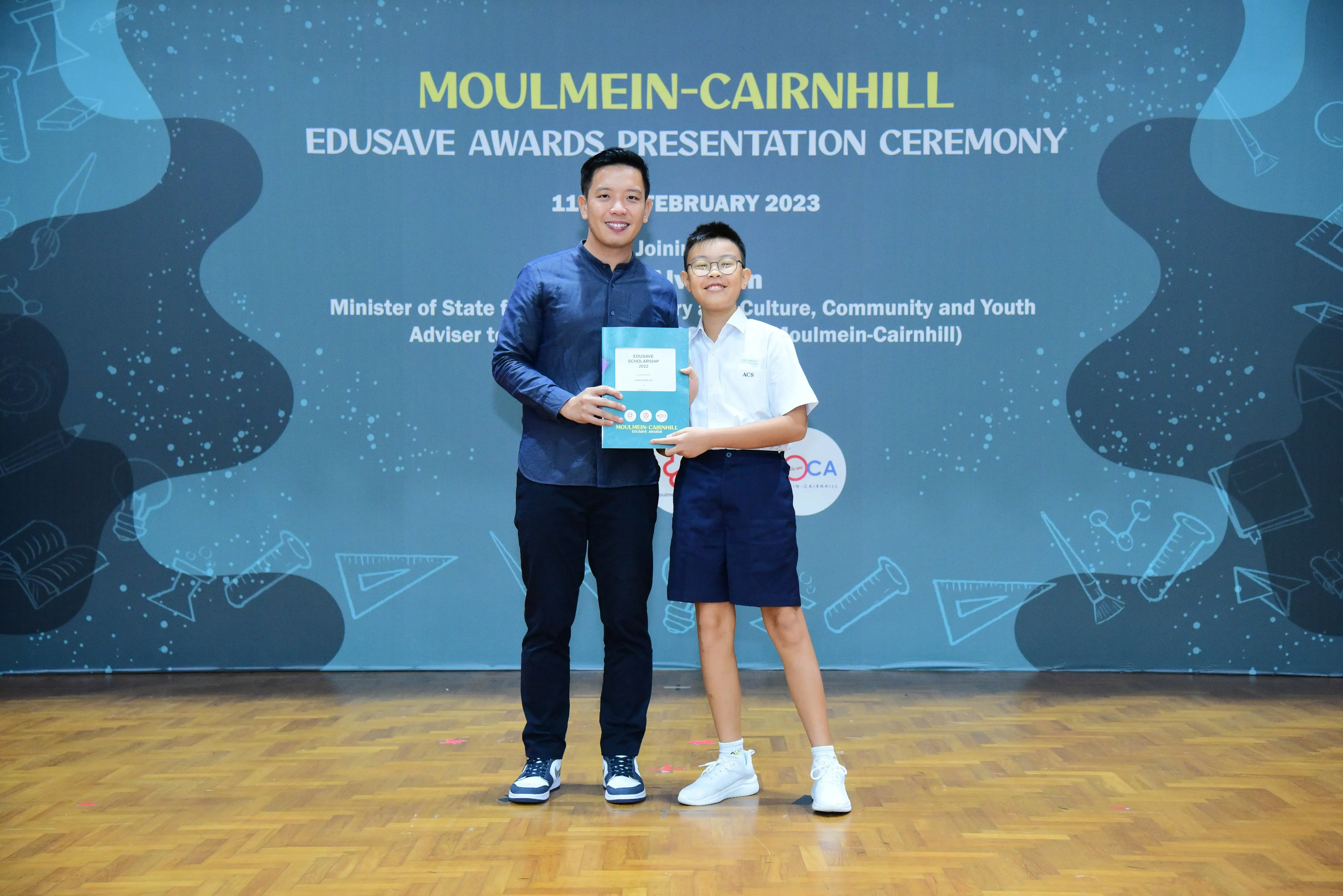 An adult and a young student hold a certificate at the Moulmein-Cairnhill Edusave Awards Presentation Ceremony 2023.