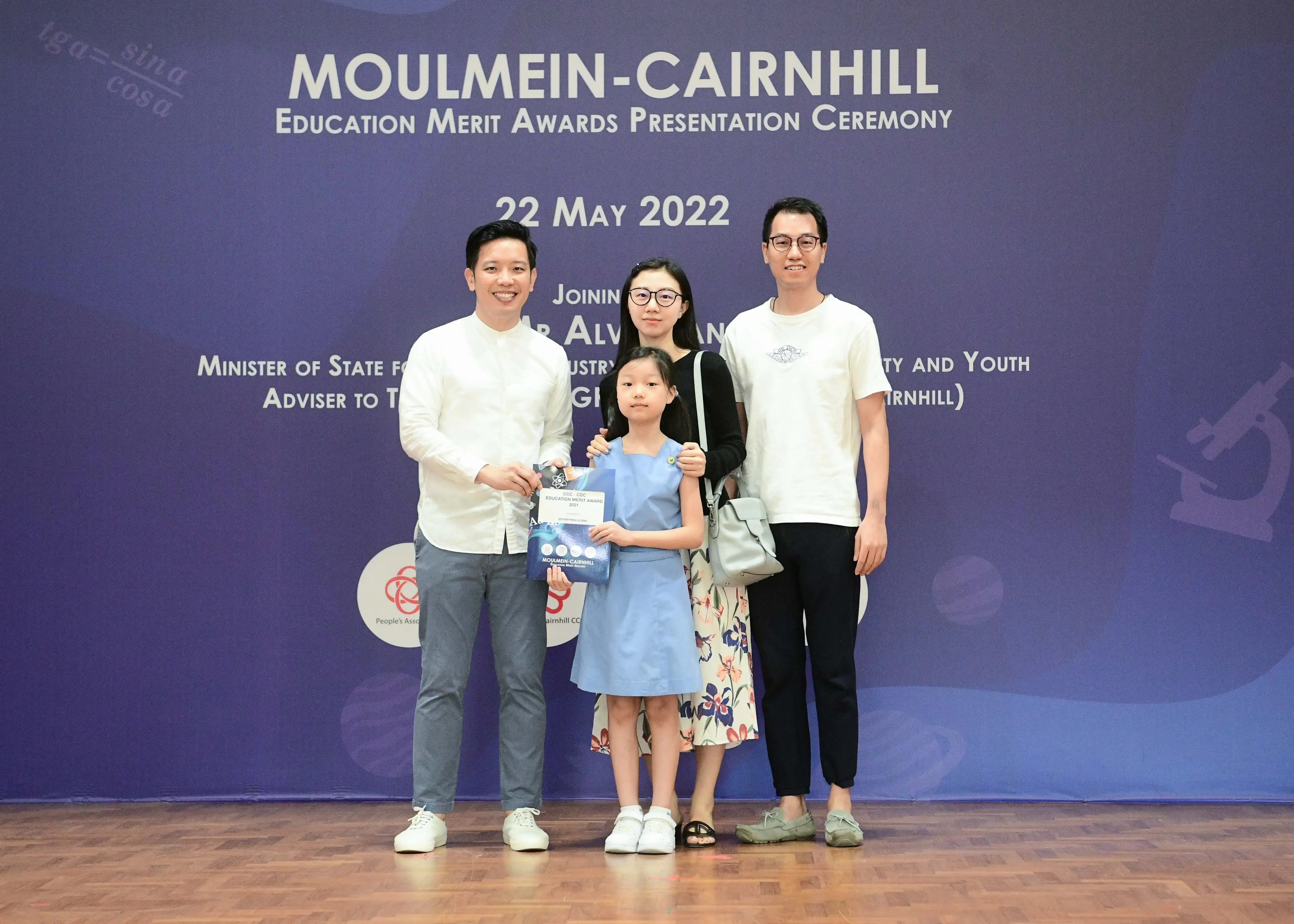 Four people pose before a Moulmein-Cairnhill Education Merit Awards banner dated 22 May 2022, with a girl holding an award.