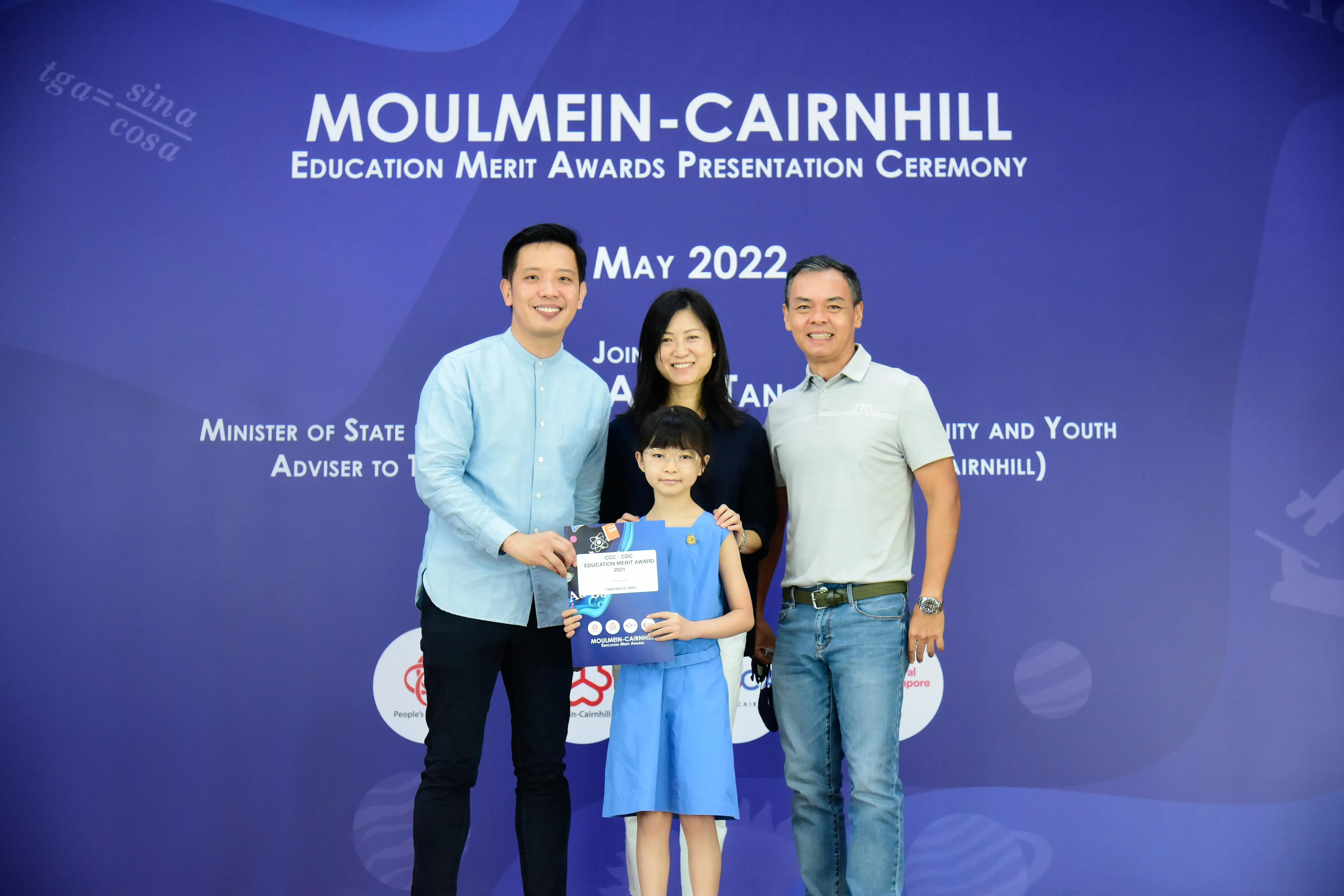 A young girl holding an award certificate poses with three adults at the Moulmein-Cairnhill Education Merit Awards May 2022.