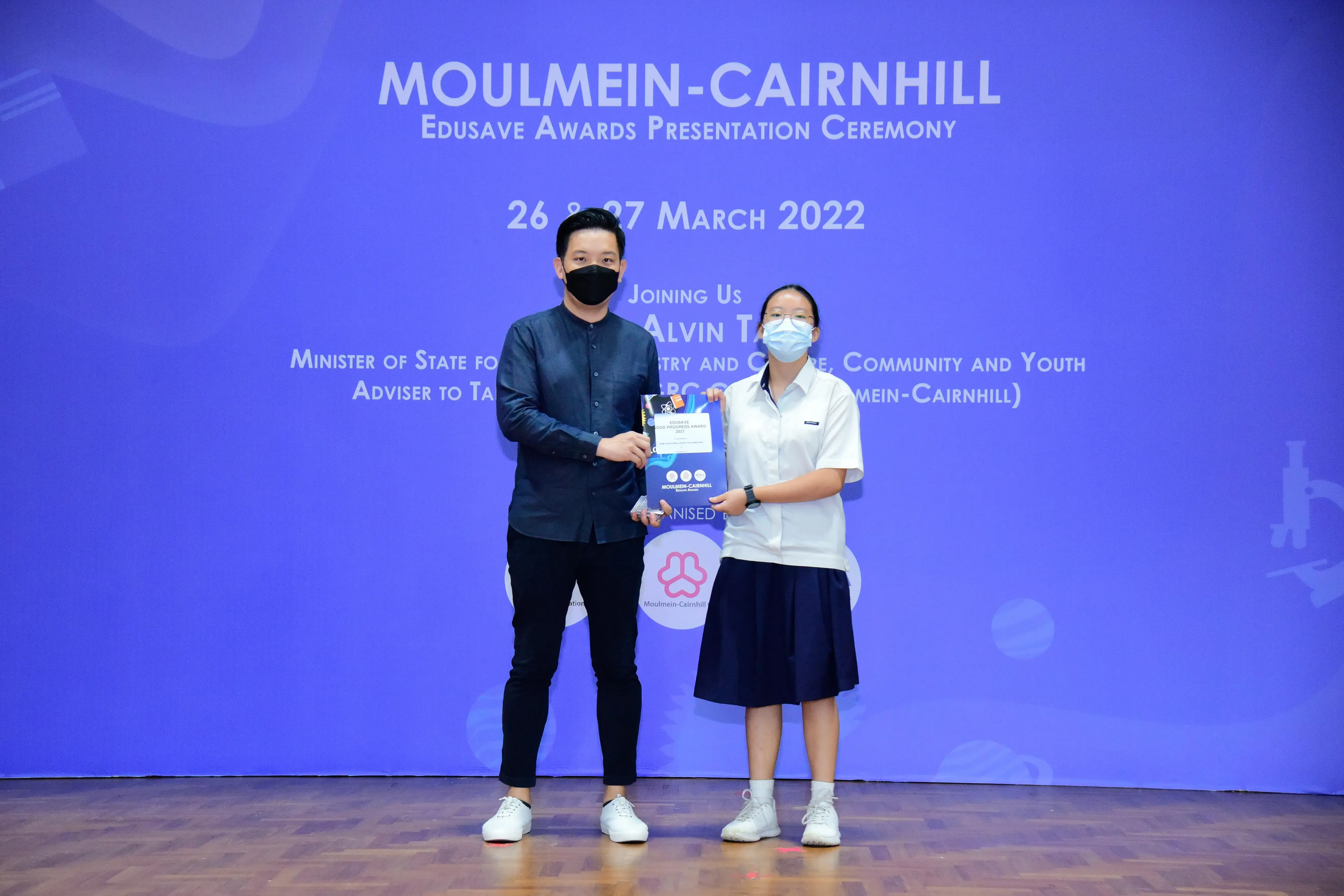 Two masked individuals hold an award certificate on stage at the Moulmein-Cairnhill Edusave Awards Ceremony, March 2022.