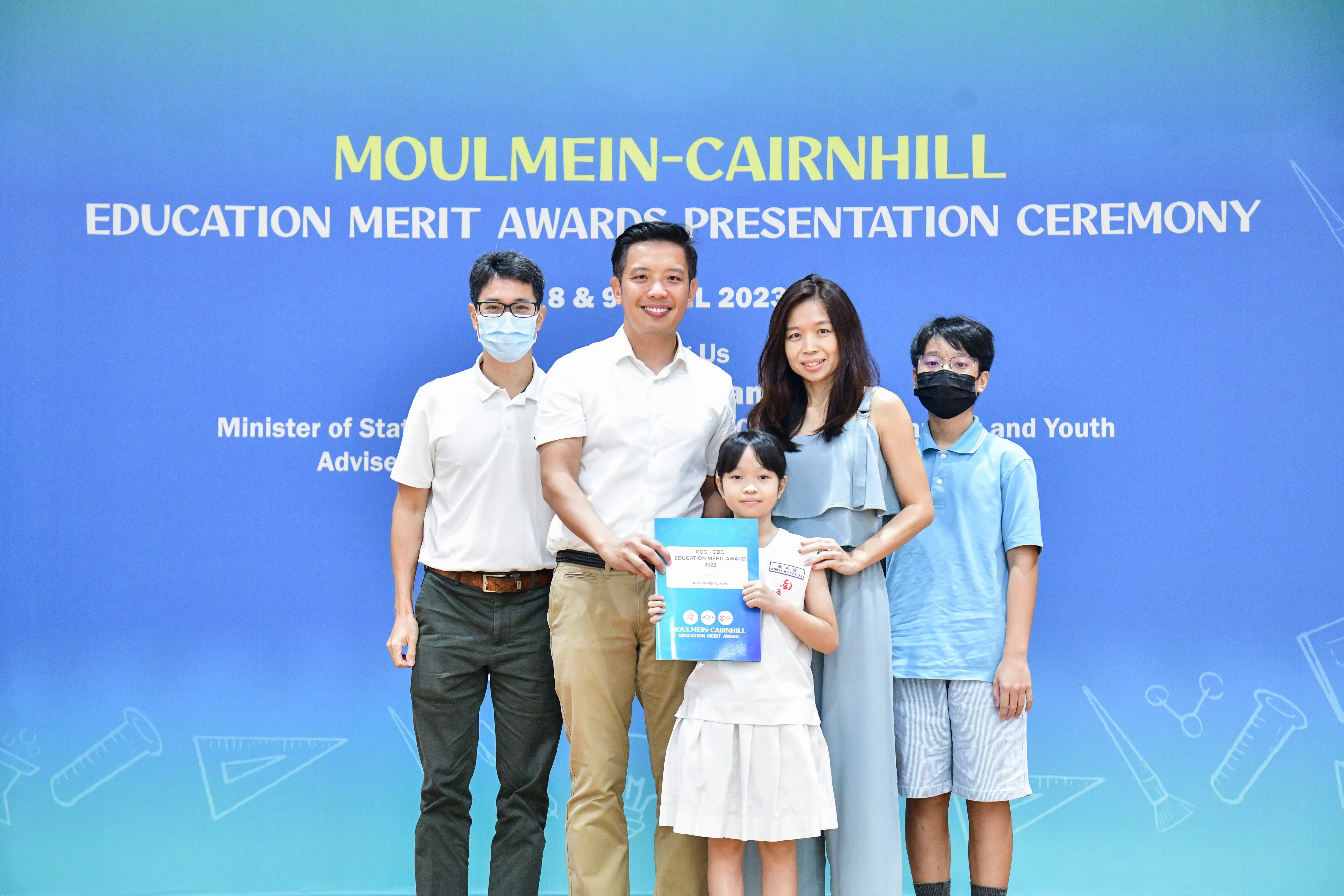 Four adults and a girl pose before a Moulmein-Cairnhill Education Merit Awards Presentation Ceremony banner in 2023.