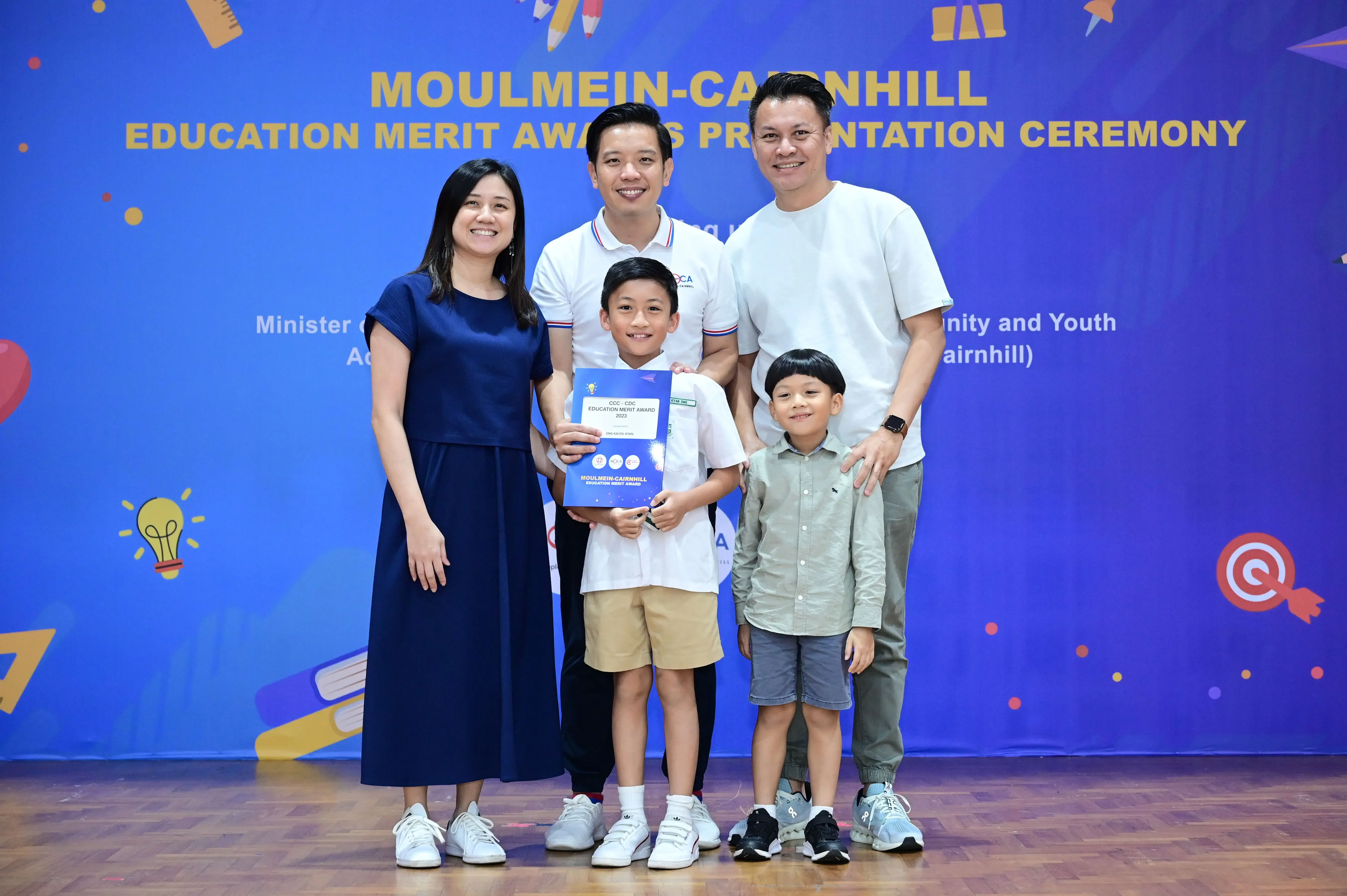 Five people pose smiling before a blue Moulmein-Cairnhill Education Merit Awards banner; a boy holds a certificate.