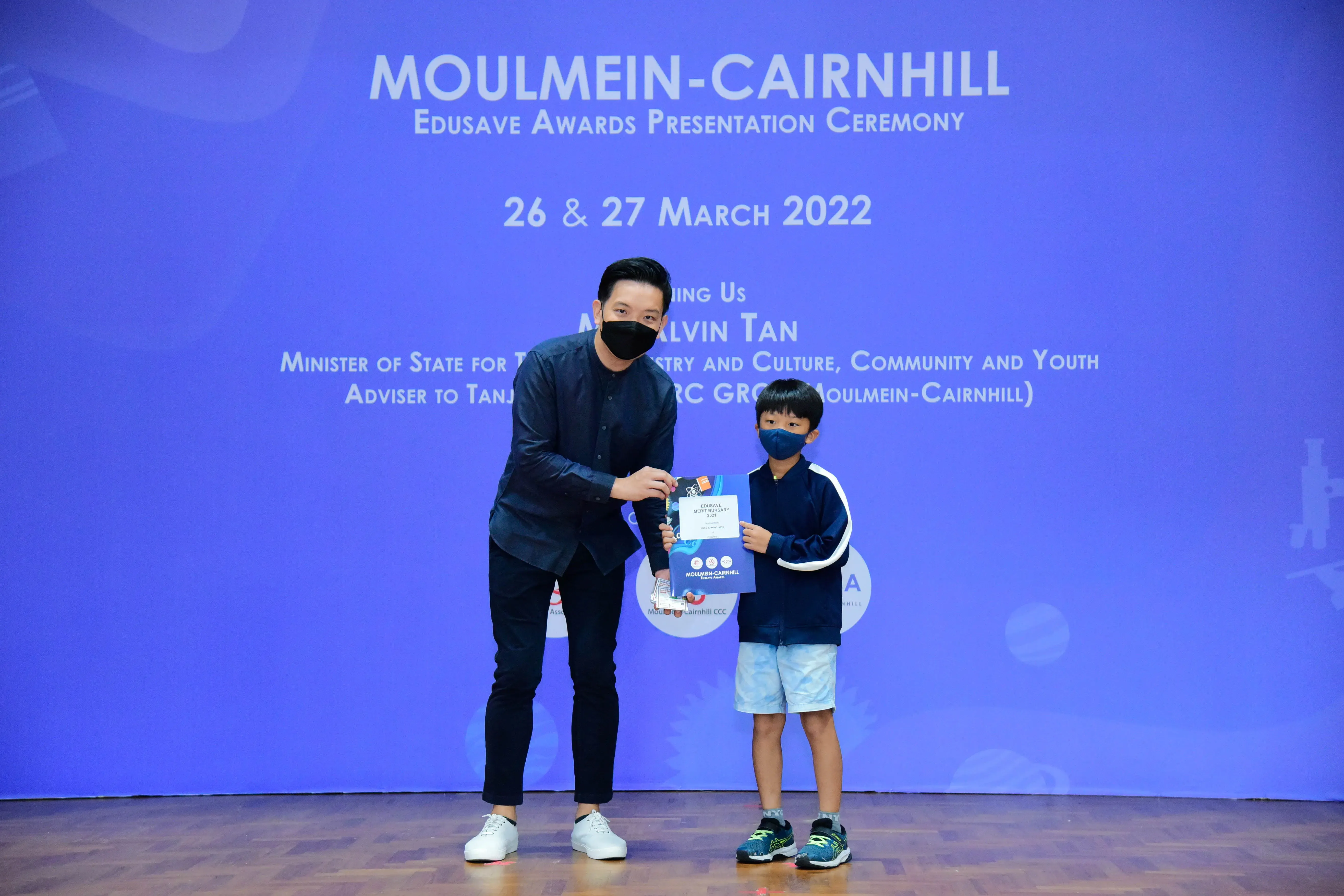 An adult and a young child in masks hold a certificate on stage at the Moulmein-Cairnhill Edusave Awards Ceremony 2022.