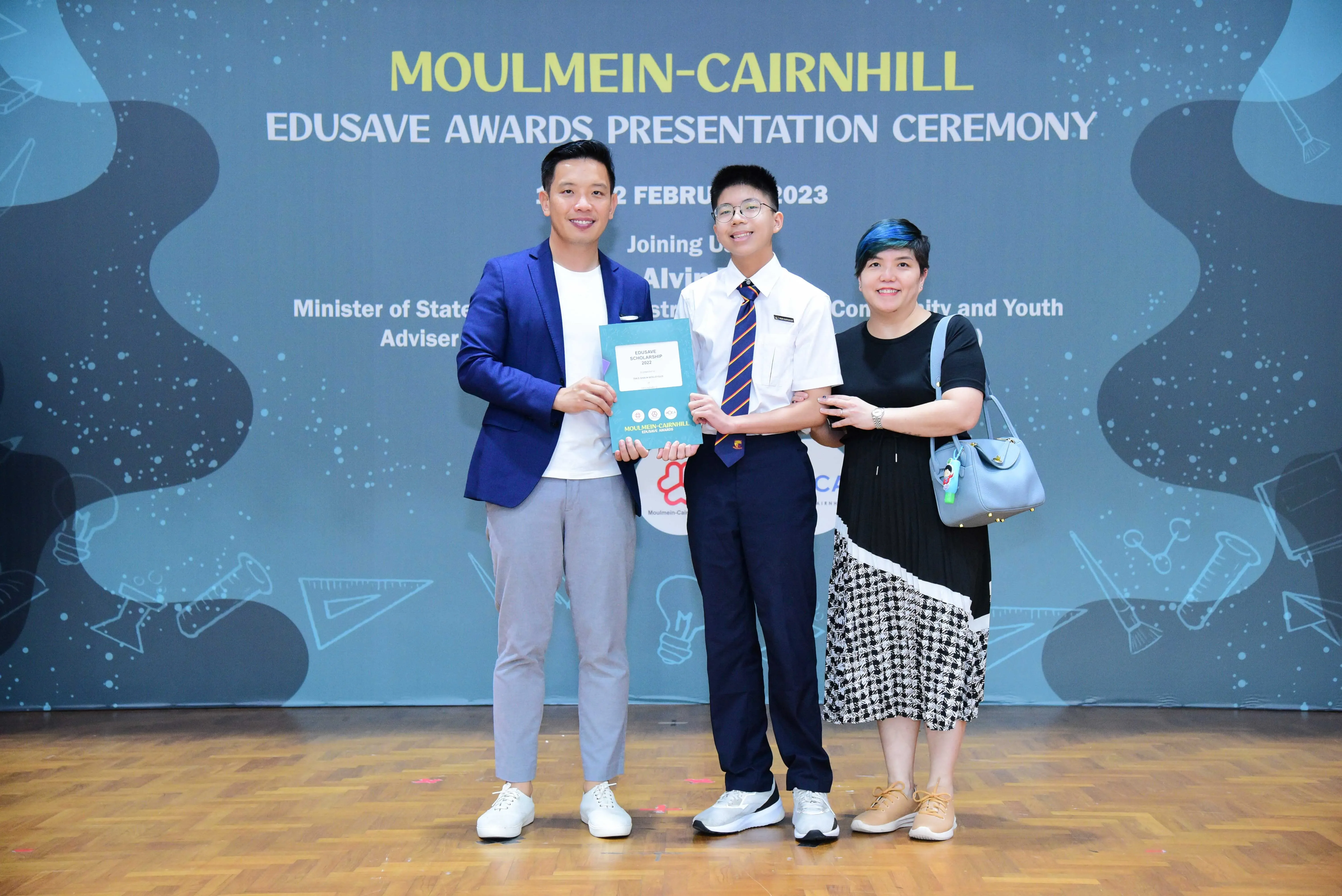 Three people smile and hold a certificate at the Moulmein-Cairnhill Edusave Awards Presentation Ceremony 2023.