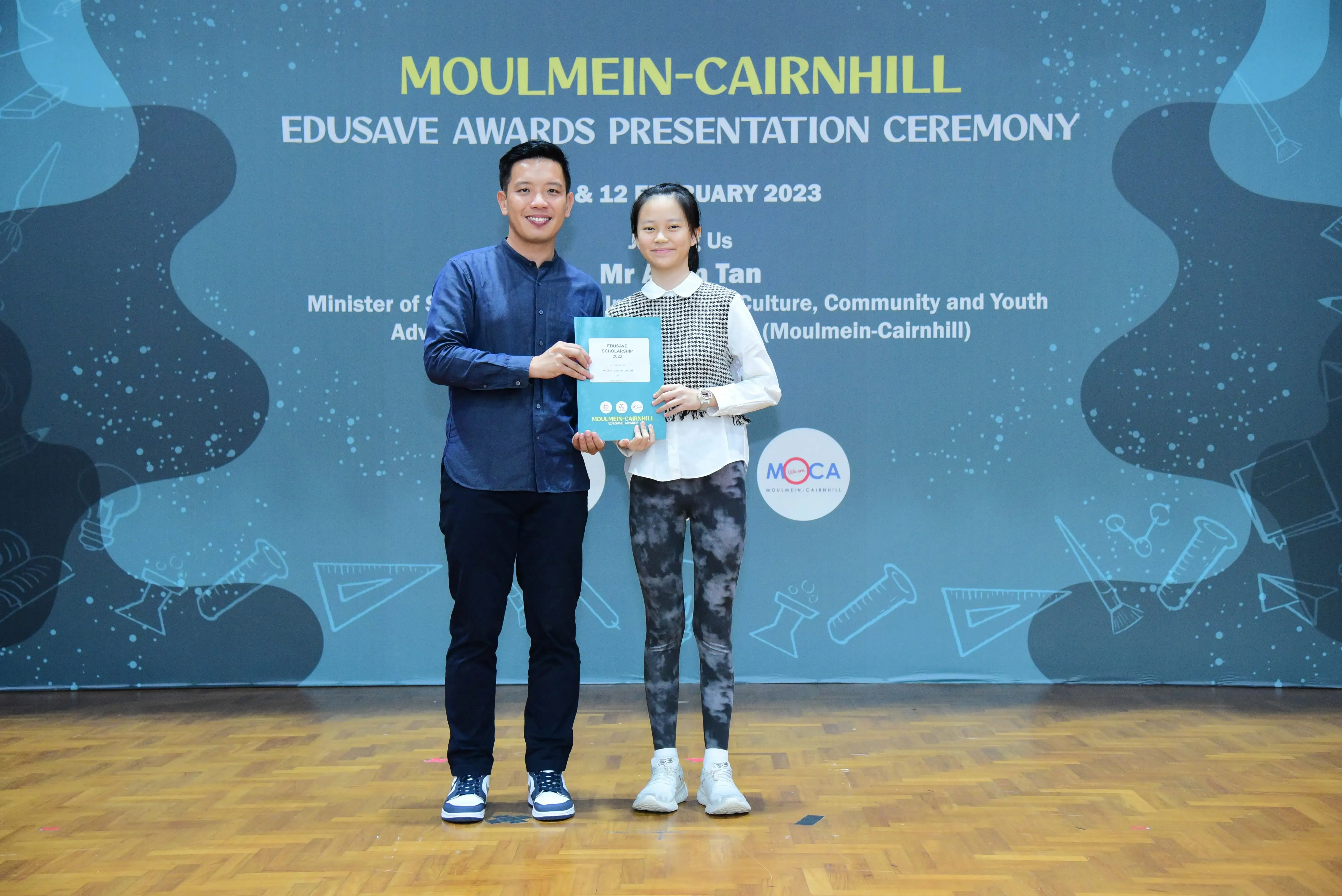 Two people smile while holding a certificate at the Moulmein-Cairnhill Edusave Awards Presentation Ceremony 2023.