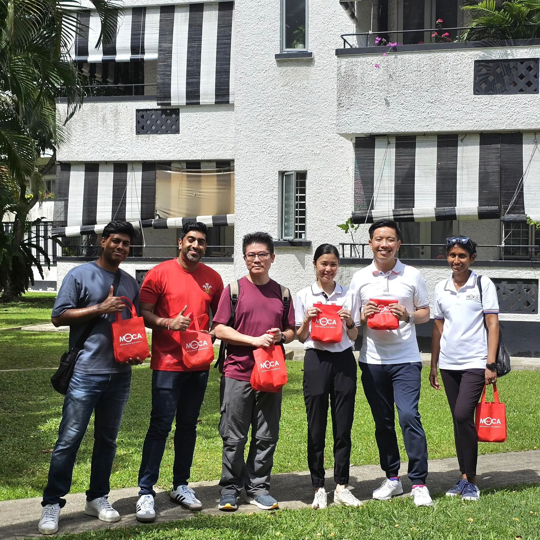 Six people stand outdoors holding small red MOCA tote bags, smiling in front of a white building with striped awnings.