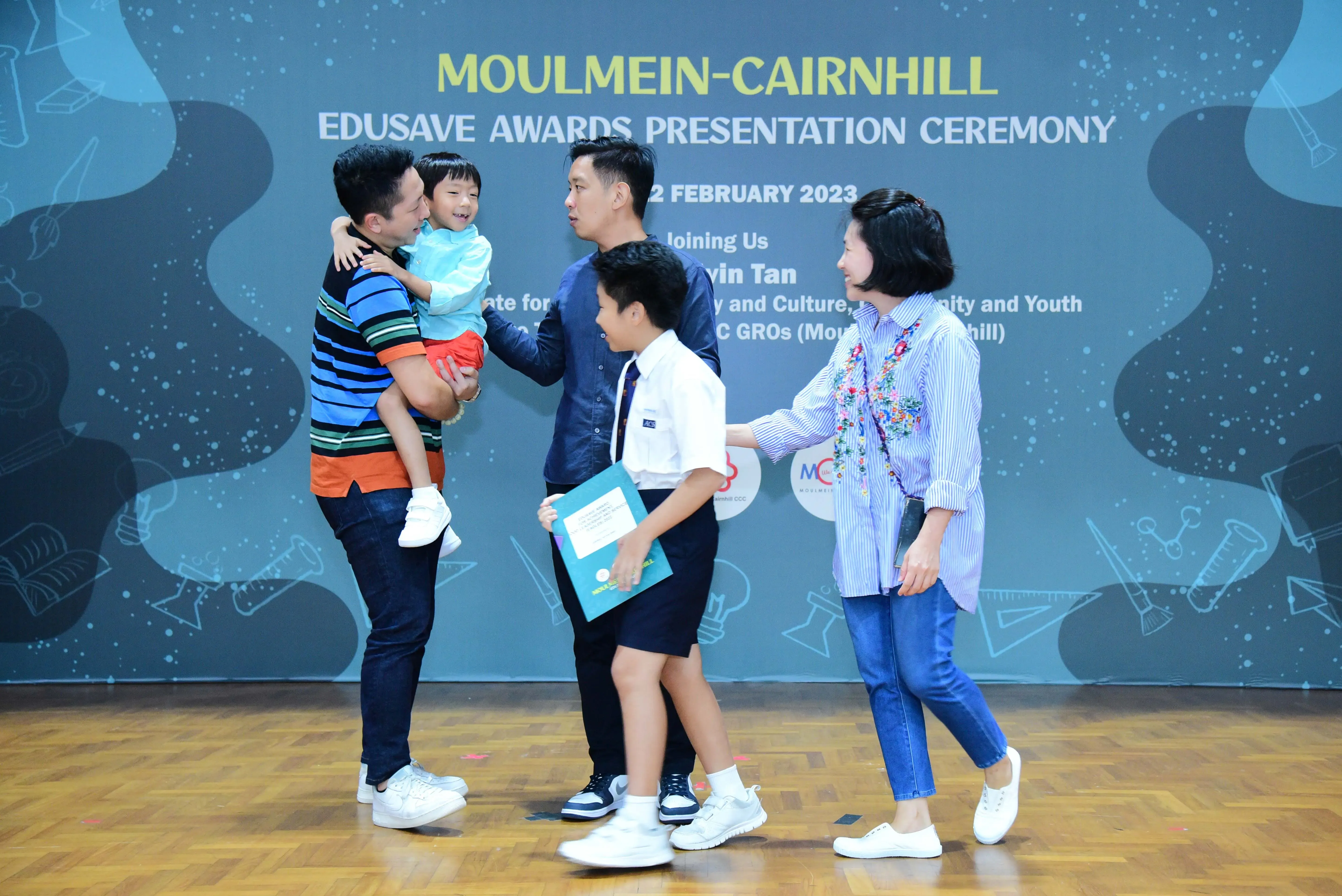 Family of four with a toddler poses before Moulmein-Cairnhill Edusave Awards Presentation Ceremony banner, February 2023.