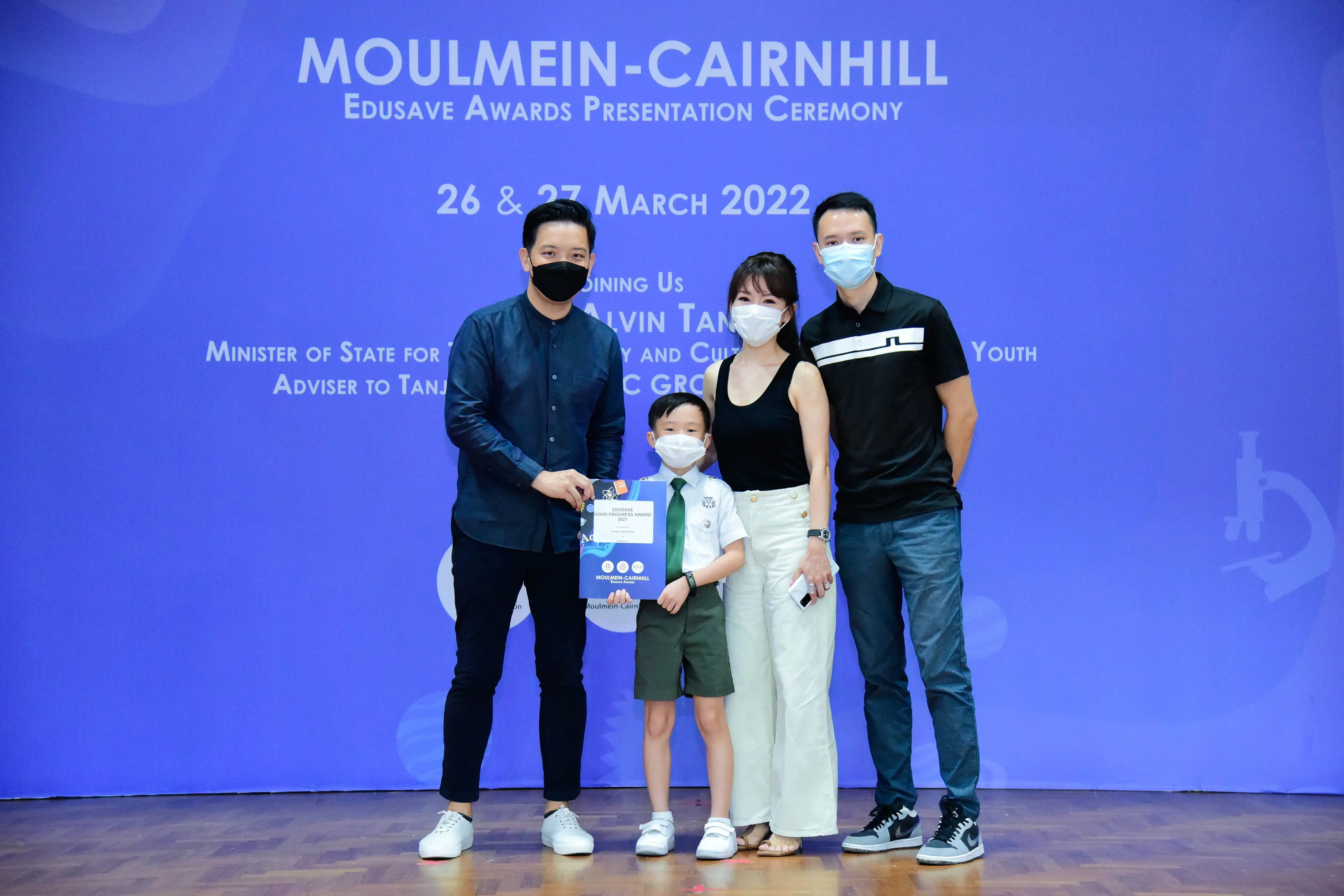 Four masked people, including a young boy in school uniform holding an award, pose at the Moulmein-Cairnhill Edusave ceremony.