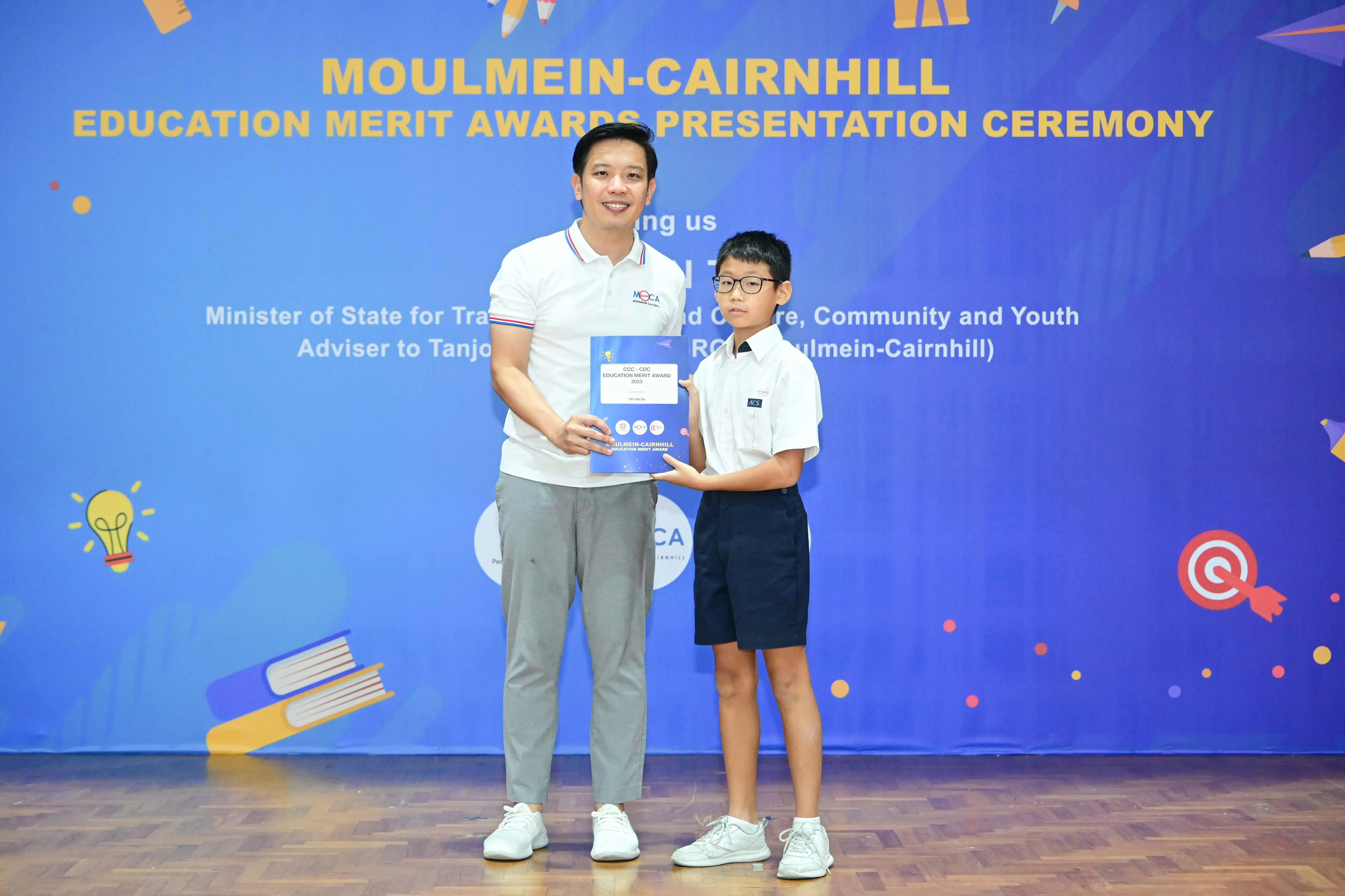 Adult and young boy hold an award booklet at Moulmein-Cairnhill Education Merit Awards Presentation Ceremony.