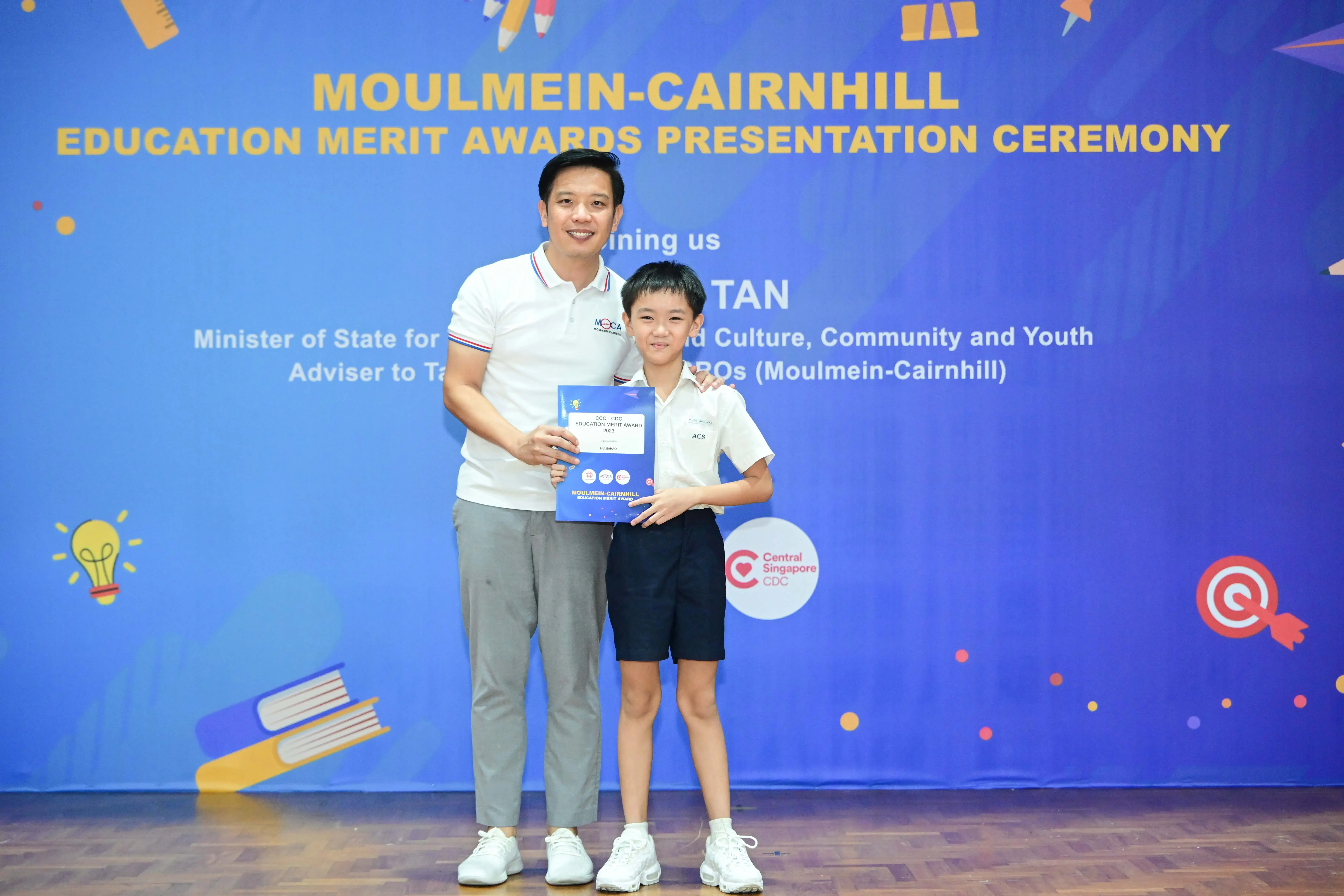 An adult and a young boy pose holding a certificate at the Moulmein-Cairnhill Education Merit Awards Ceremony.