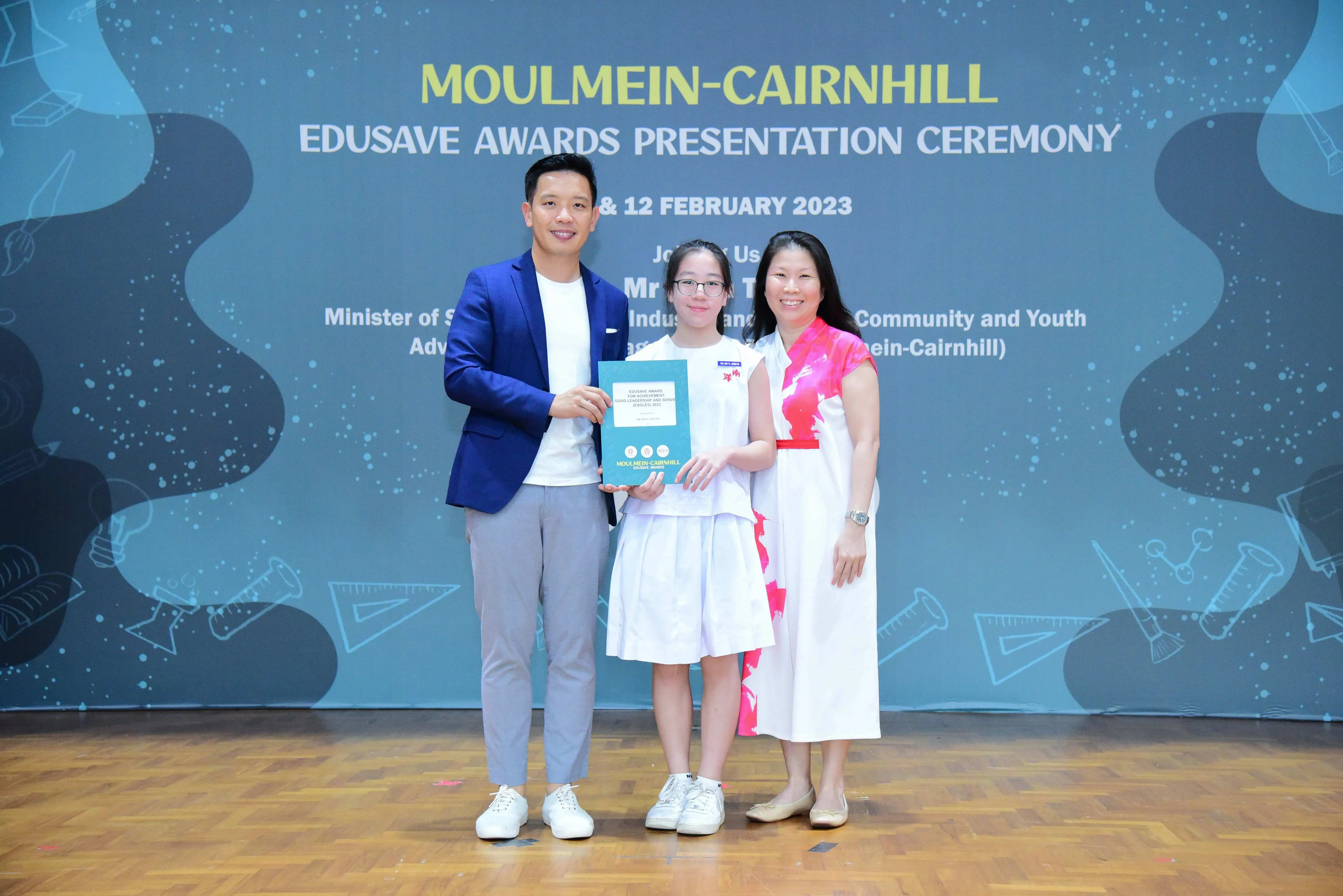 Three people pose on a stage at the Moulmein-Cairnhill Edusave Awards Presentation Ceremony, holding a teal award booklet.