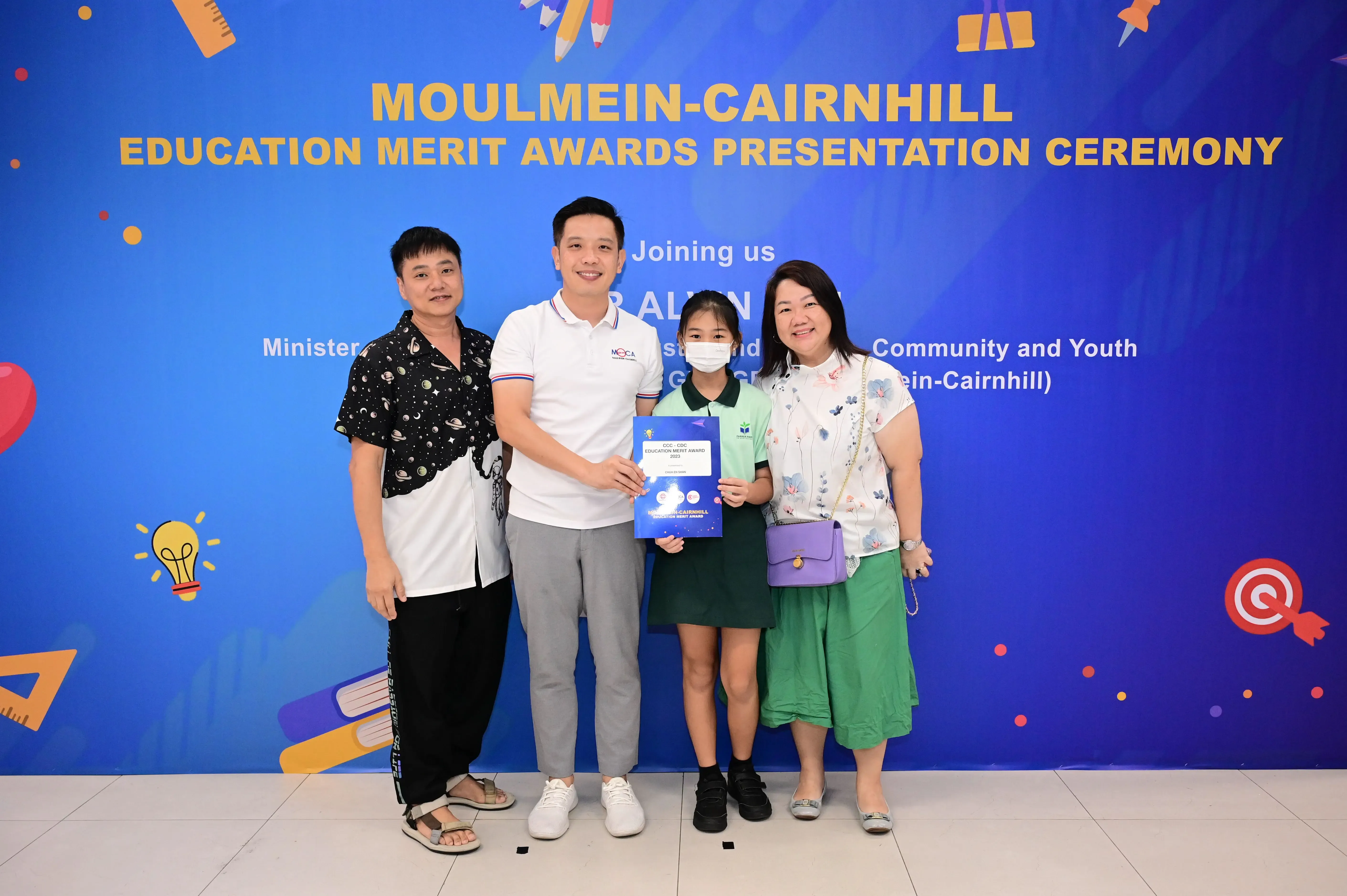Four people pose before a Moulmein-Cairnhill Education Merit Awards banner, with a girl holding a blue certificate.