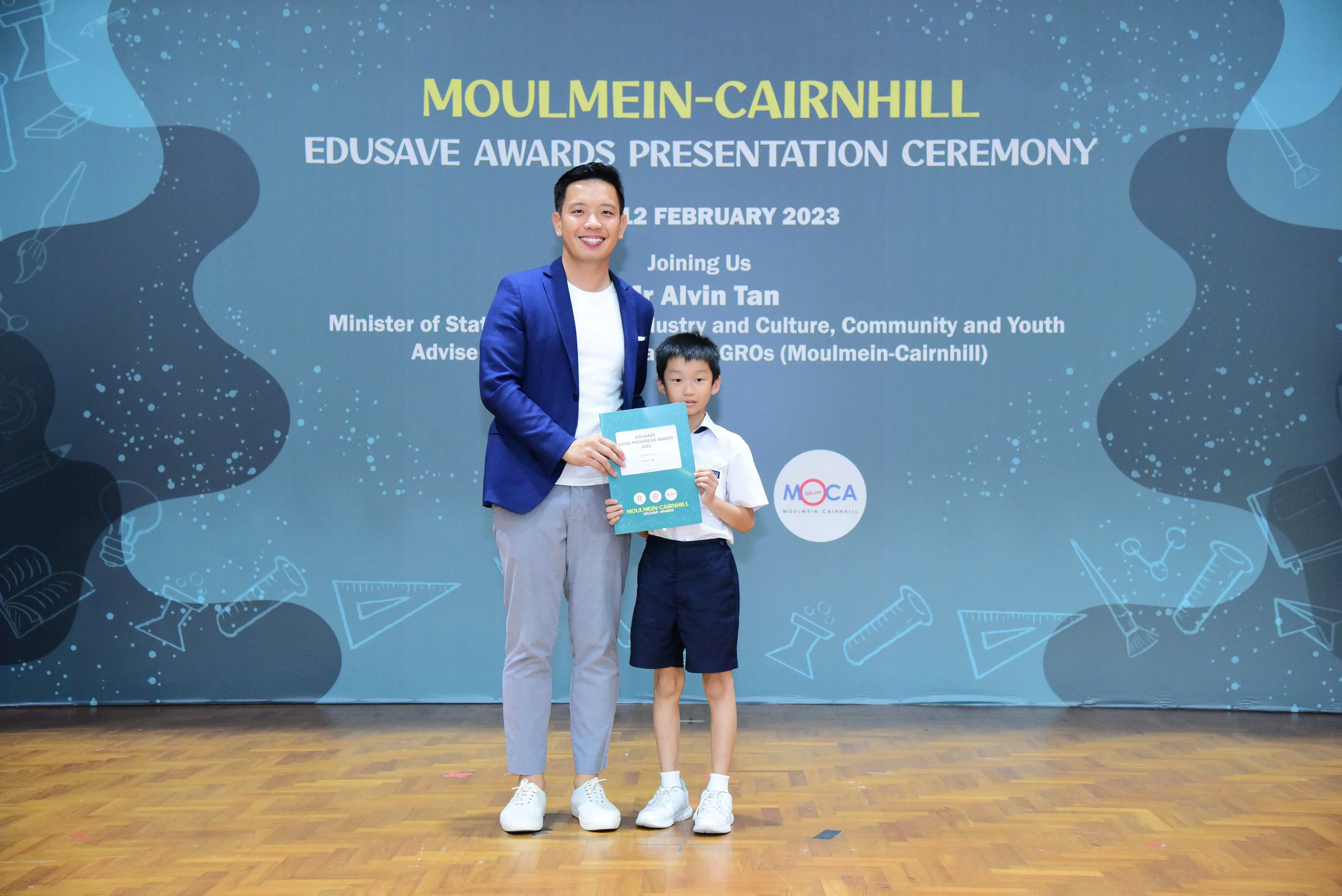 A man in a blue blazer and a young boy in school uniform hold a certificate at the Moulmein-Cairnhill Edusave Awards 2023.