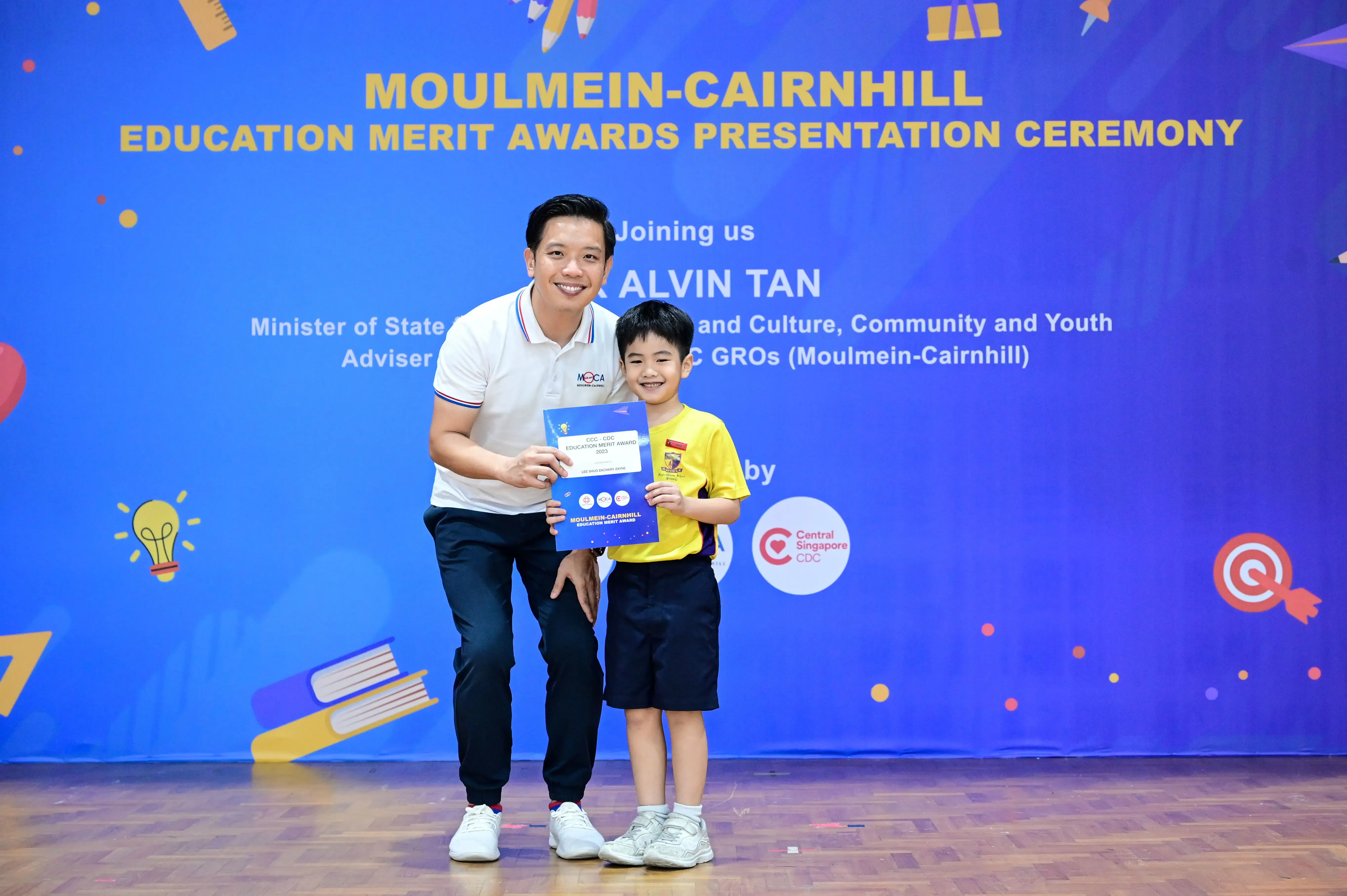 A man and a young boy hold a certificate together in front of a Moulmein-Cairnhill Education Merit Awards banner.