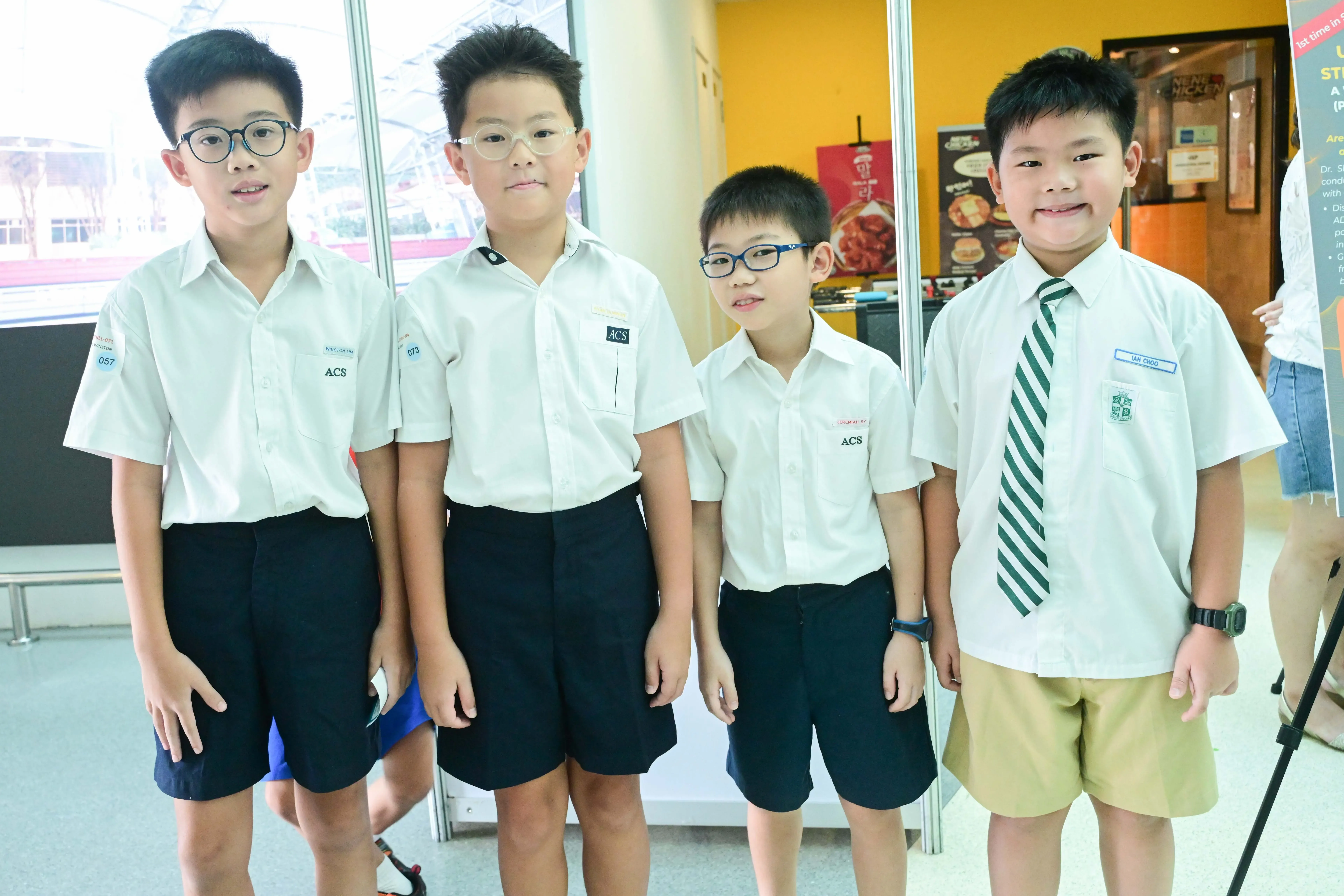 Four young boys in ACS school uniforms stand together, three wearing glasses, one with a green striped tie.
