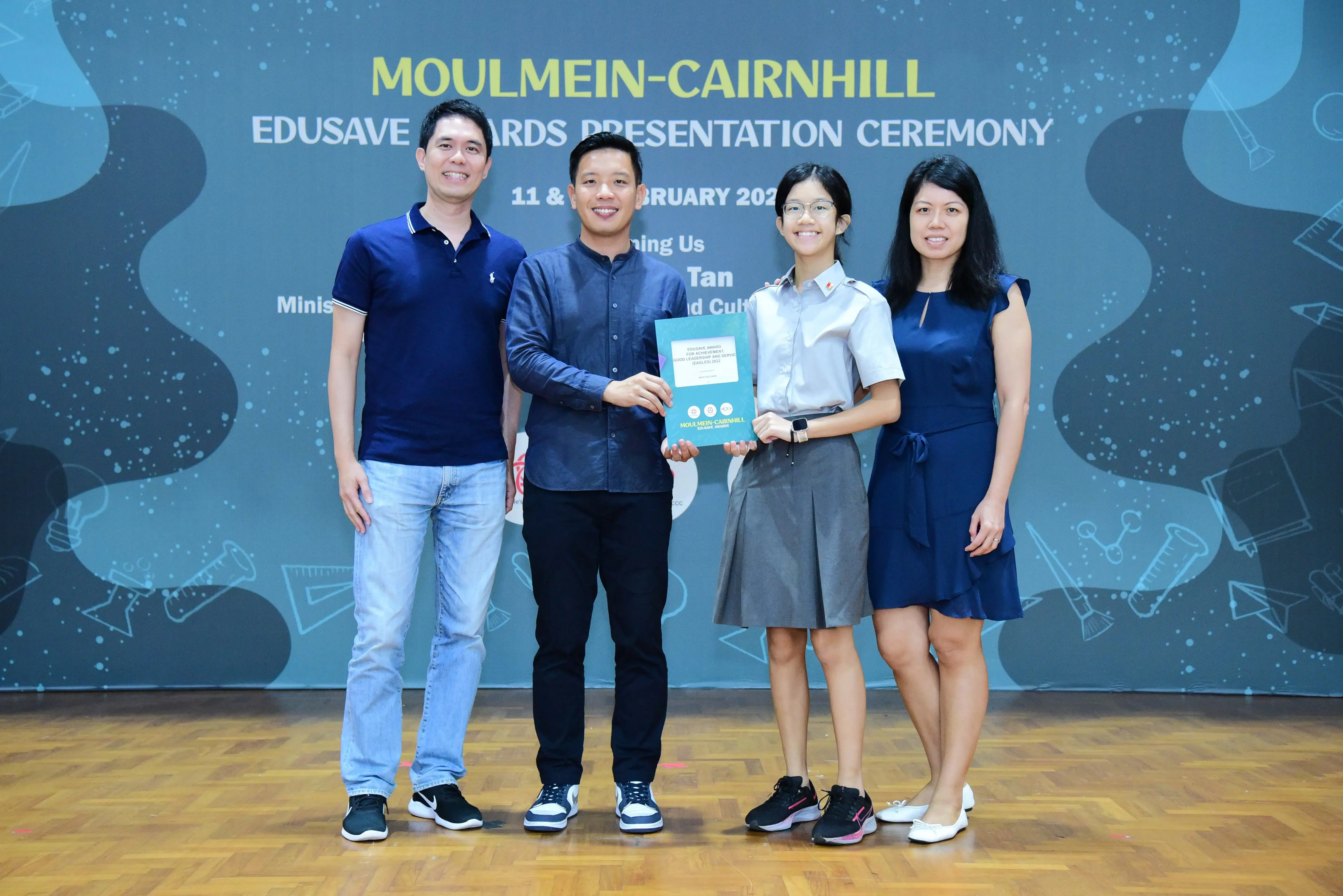Four people smiling on stage at the Moulmein-Cairnhill Edusave Awards Presentation Ceremony, one holding a certificate.