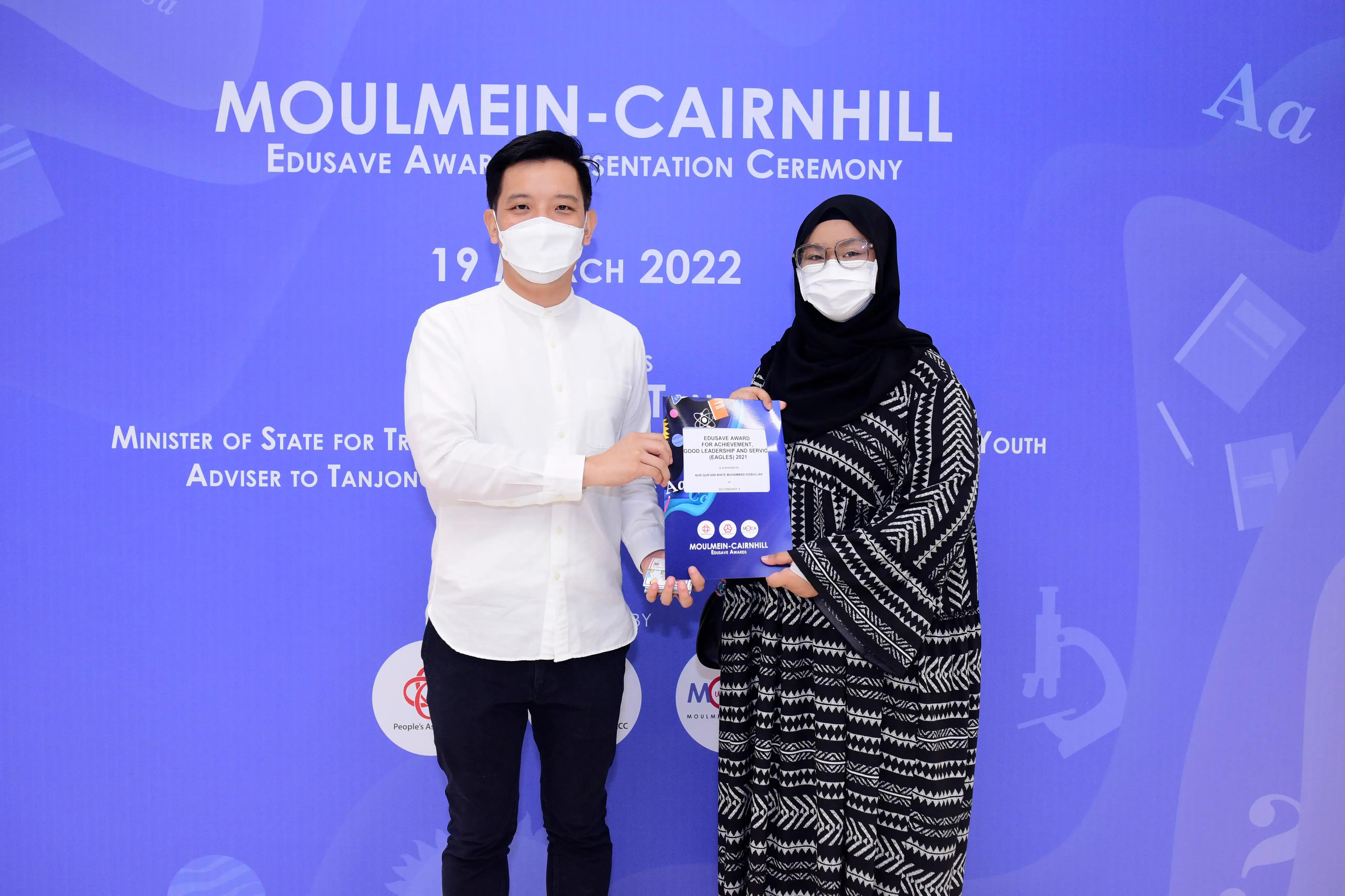 Two masked people hold a certificate at the Moulmein-Cairnhill Edusave Award Presentation Ceremony on 19 March 2022.