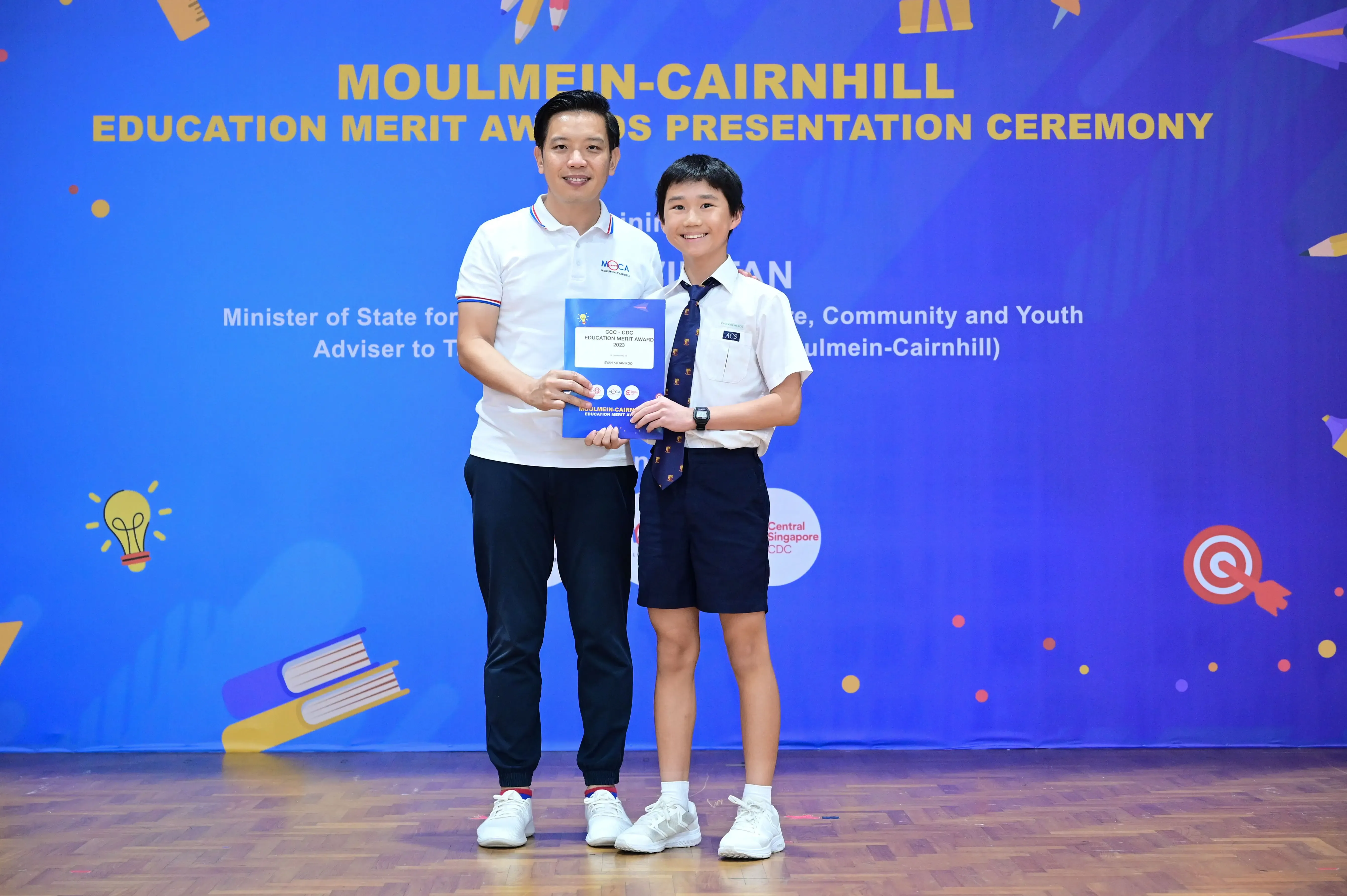 An adult and a uniformed student hold an award certificate at the Moulmein-Cairnhill Education Merit Awards Presentation Ceremony.